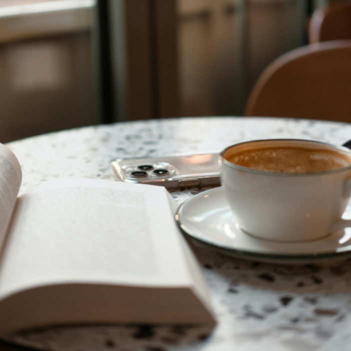 A coffee cup with a saucer, an open book, and a smartphone on a speckled table in a cozy café setting.