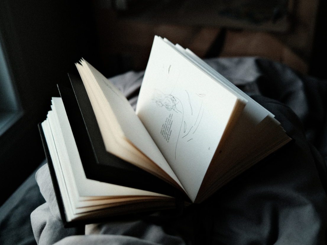 An open sketchbook with a black cover resting on a dark surface, with a simple line drawing of a woman's face and some handwritten text, with blurred background.