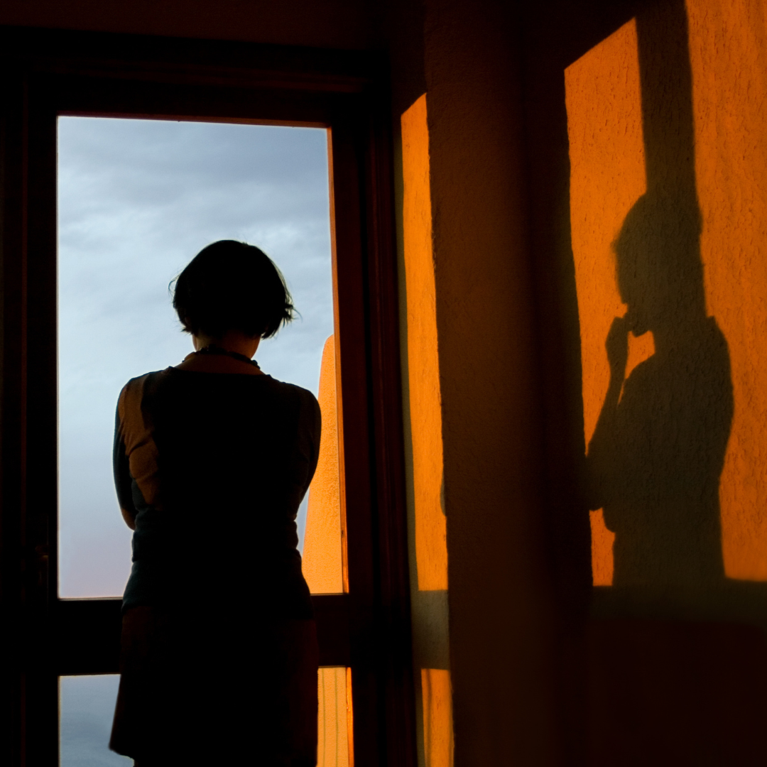 Silhouette of a person standing near a window with an orange wall beside it, with their shadow cast on the wall, and a cloudy sky visible outside.