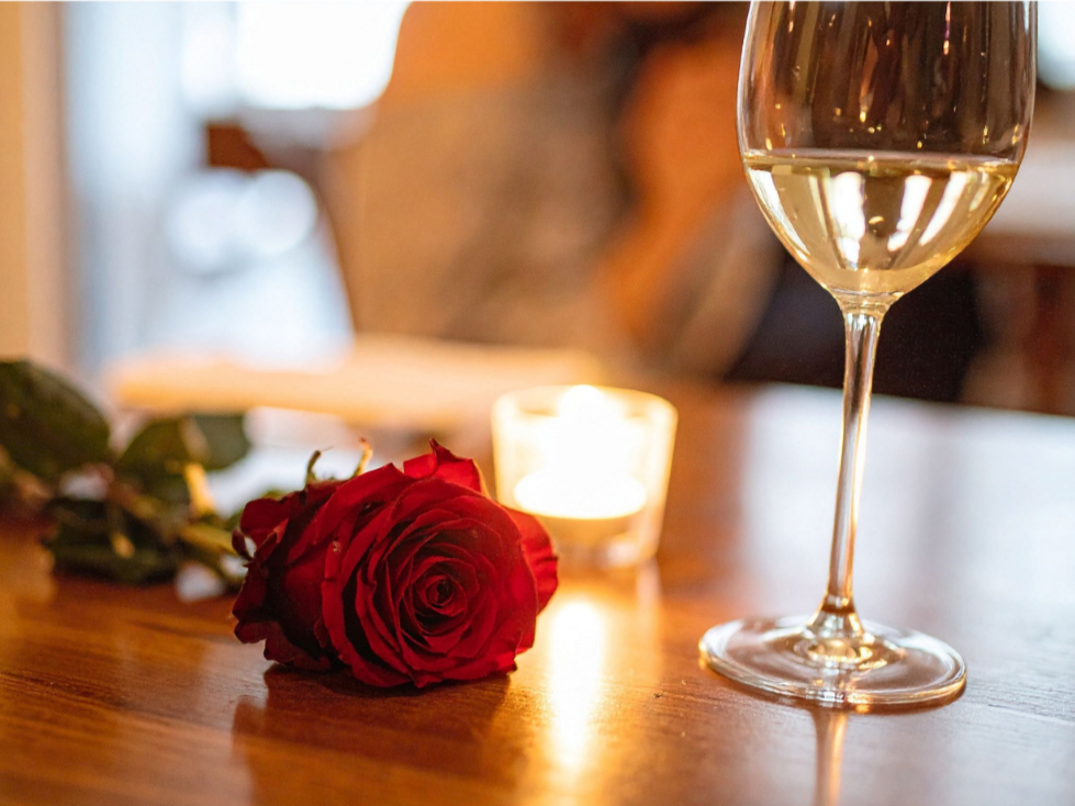 A red rose, a lit candle, and a glass of white wine on a wooden table.