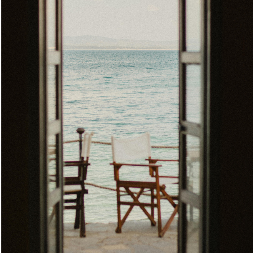 View of the ocean through an open door with a wooden chair and a small table on a balcony or porch.