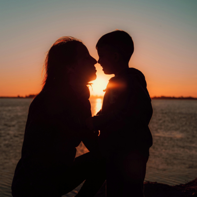 Silhouette of a woman and a young boy facing each other, touching noses, during sunset at a body of water.