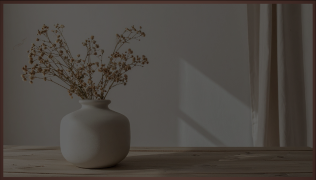 A white vase with dried flowers on a wooden surface in a well-lit room.