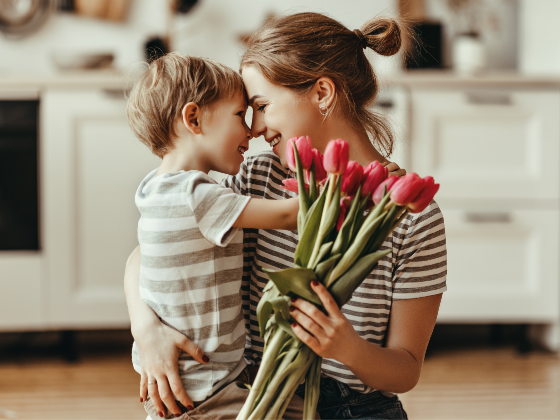 A woman and a young boy are touching their foreheads together and smiling in a kitchen, with the woman holding a bouquet of pink tulips.