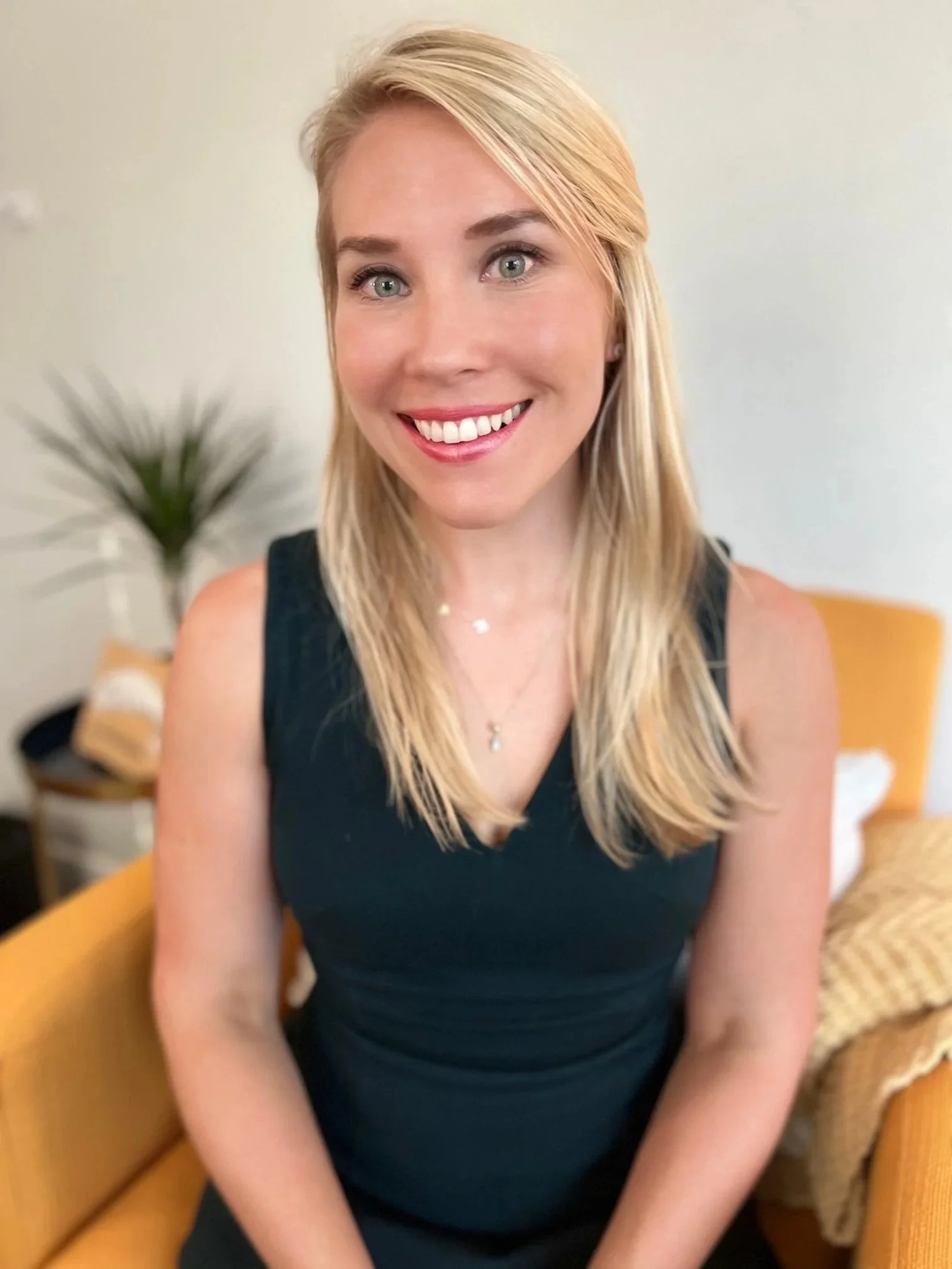 A smiling woman with blonde hair wearing a black sleeveless top, sitting on a yellow chair in a room with a blurred background that includes a plant and furniture.