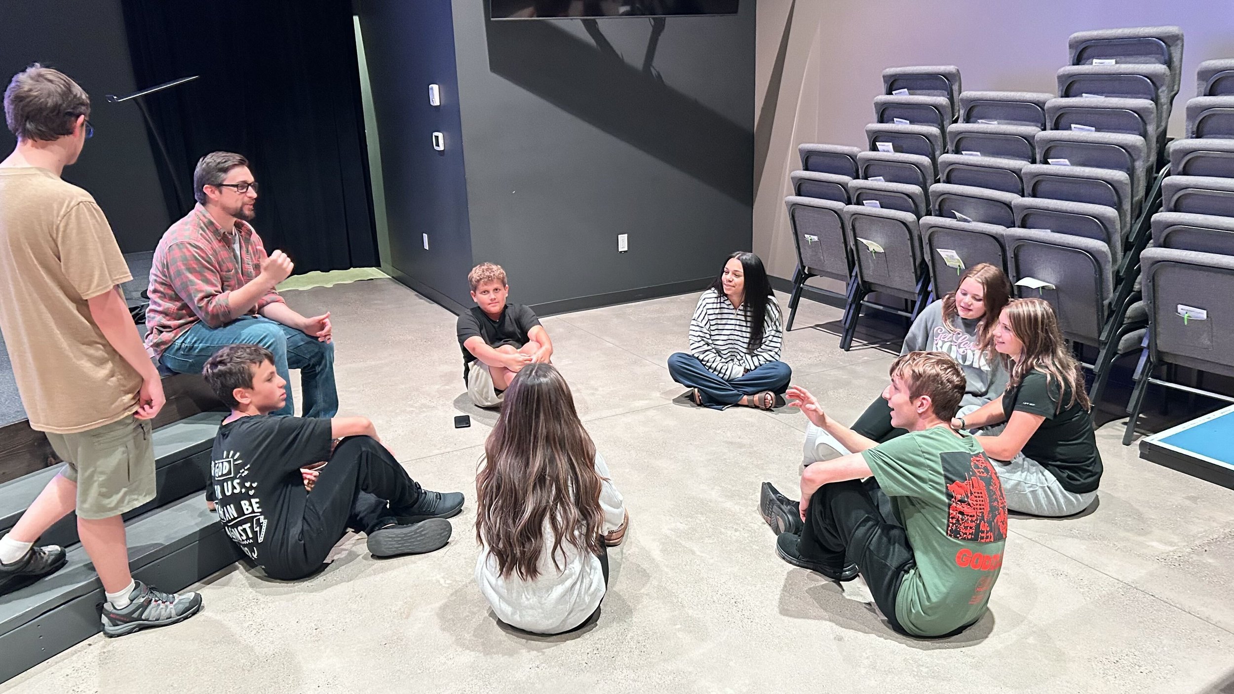 A group of young people sitting on the floor in a circle, engaged in a discussion or activity, in an indoor space with stacked chairs in the background.