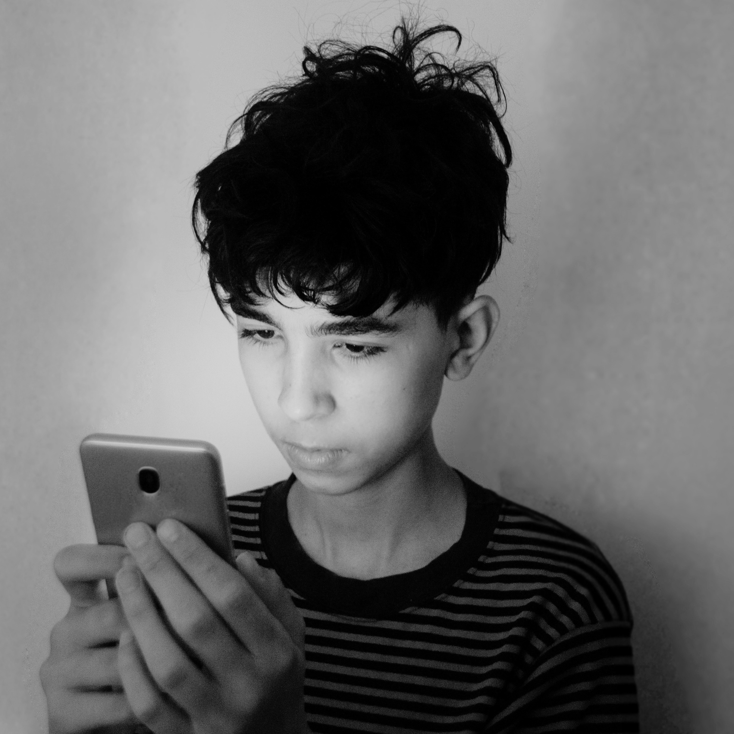 Black and white photo of a young man with curly hair looking down at a phone. He is wearing a striped shirt and standing against a plain background.