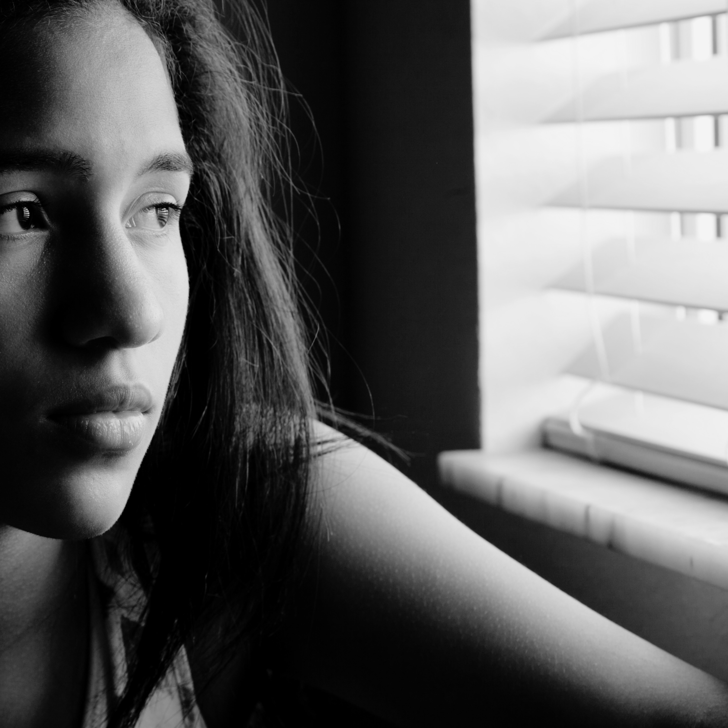 Close-up black and white photo of a woman's face with focus on her left eye and part of her forehead, with curtain blinds in the background.