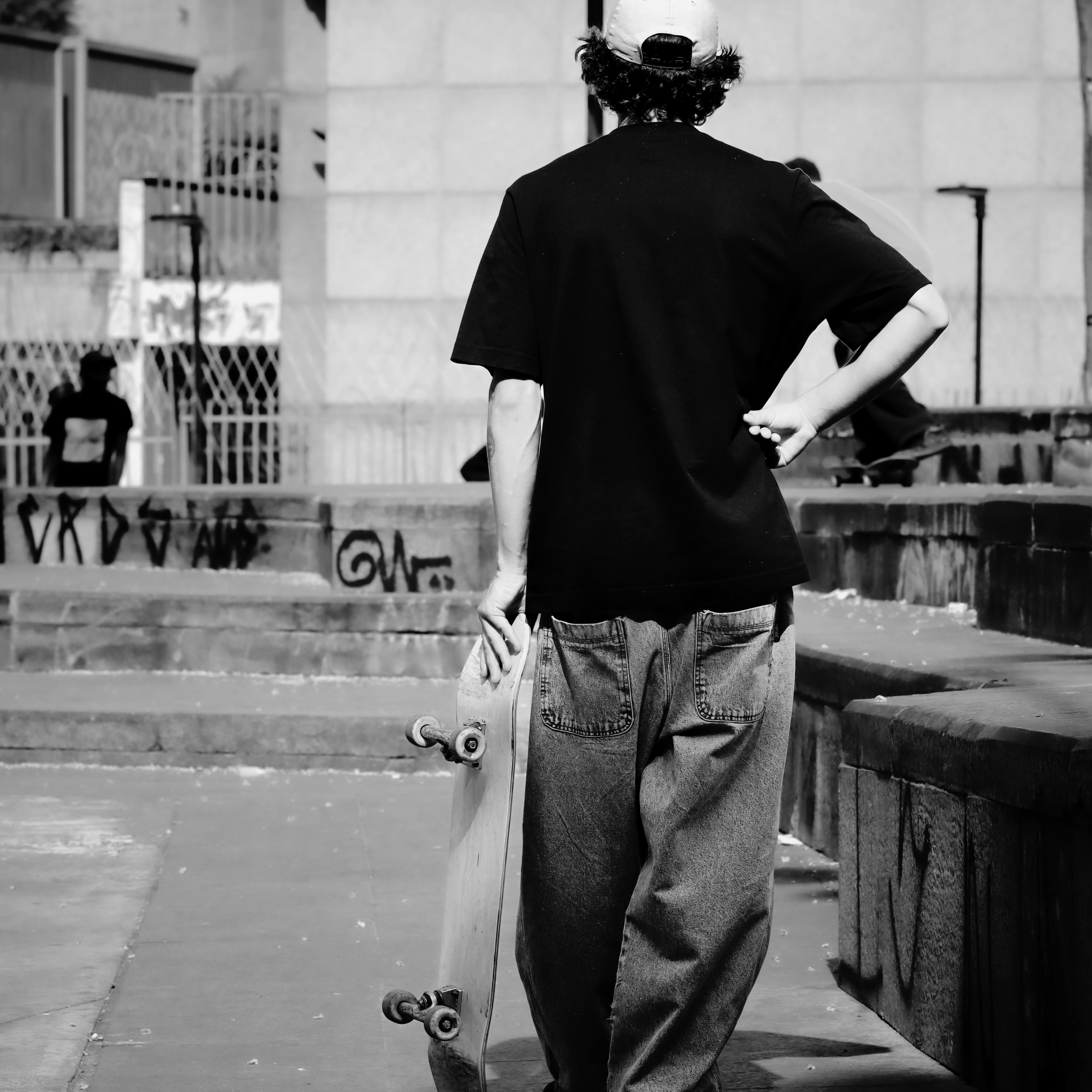 A young person standing with their back to the camera, holding a skateboard in their right hand, wearing a baseball cap and jeans, on an urban sidewalk with a building in the background, and graffiti on the ground.