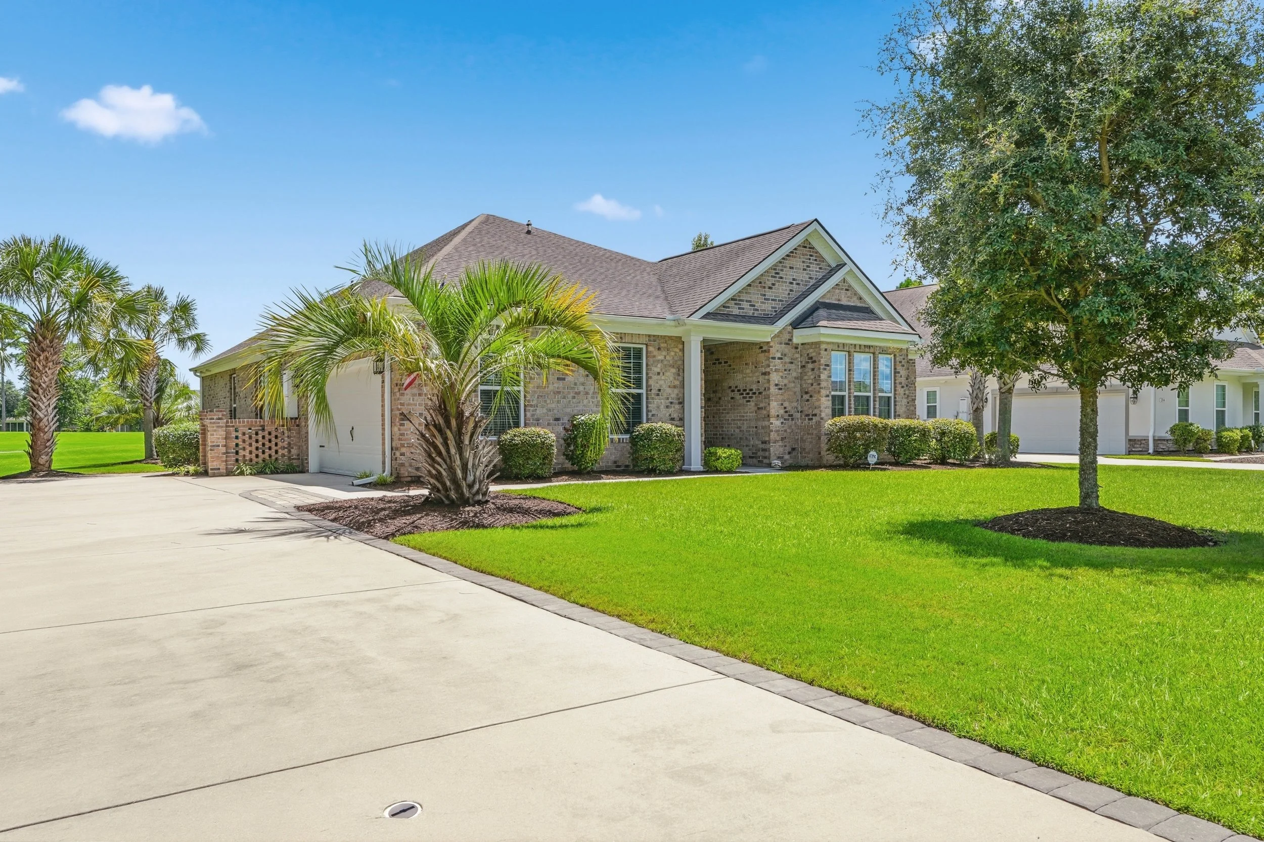 A suburban house with a brick exterior, large windows, a sloped roof, surrounded by a green lawn, trees, and palm trees, under a blue sky with a few clouds.