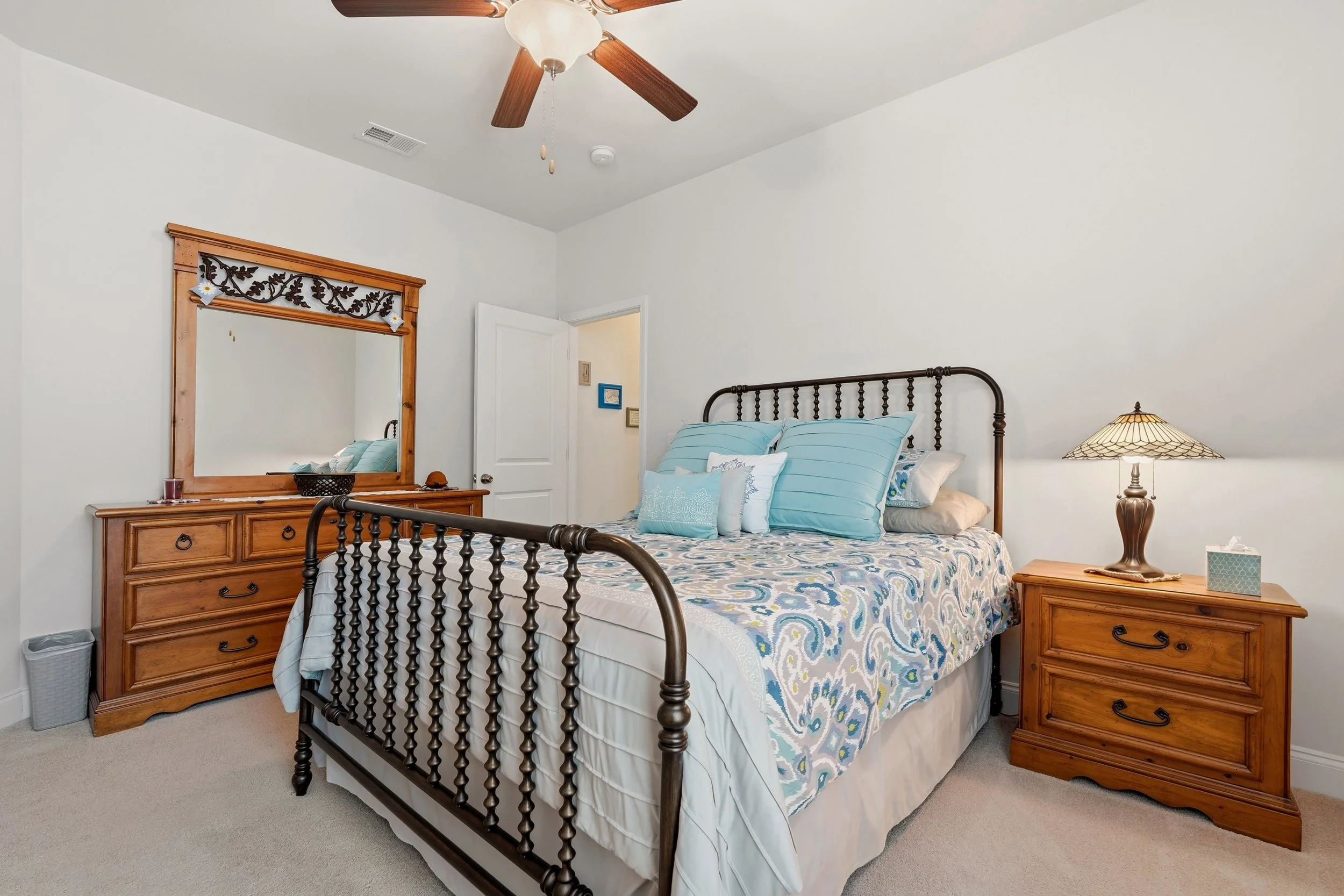 A bedroom with a metal bed, wooden nightstand, dresser with mirror, and a ceiling fan with wooden blades.