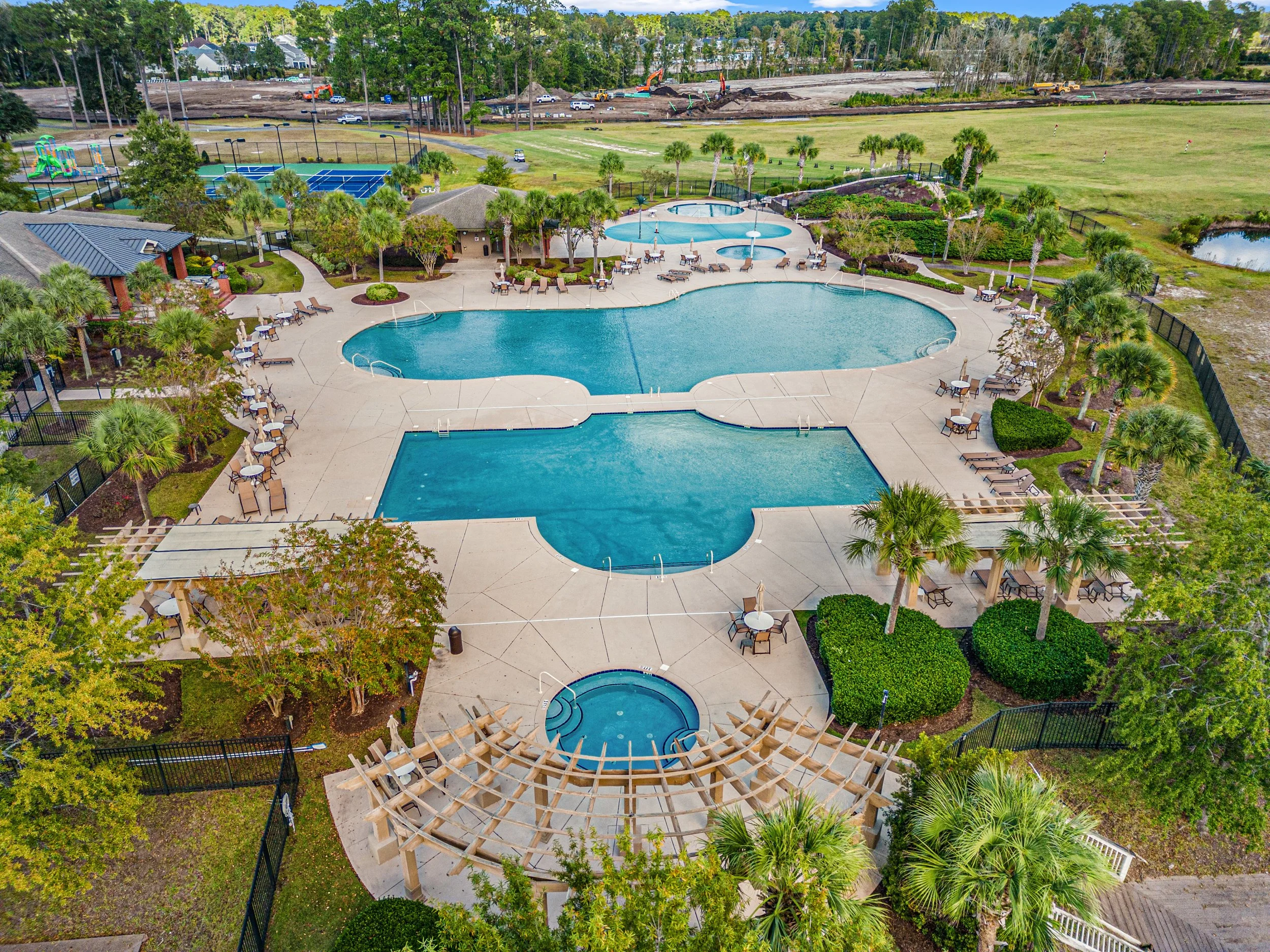 An aerial view of a large outdoor swimming pool area with lounge chairs, umbrellas, and a hot tub, surrounded by palm trees and greenery. In the background, there is a playground, and beyond that, a field and an area under construction with machinery