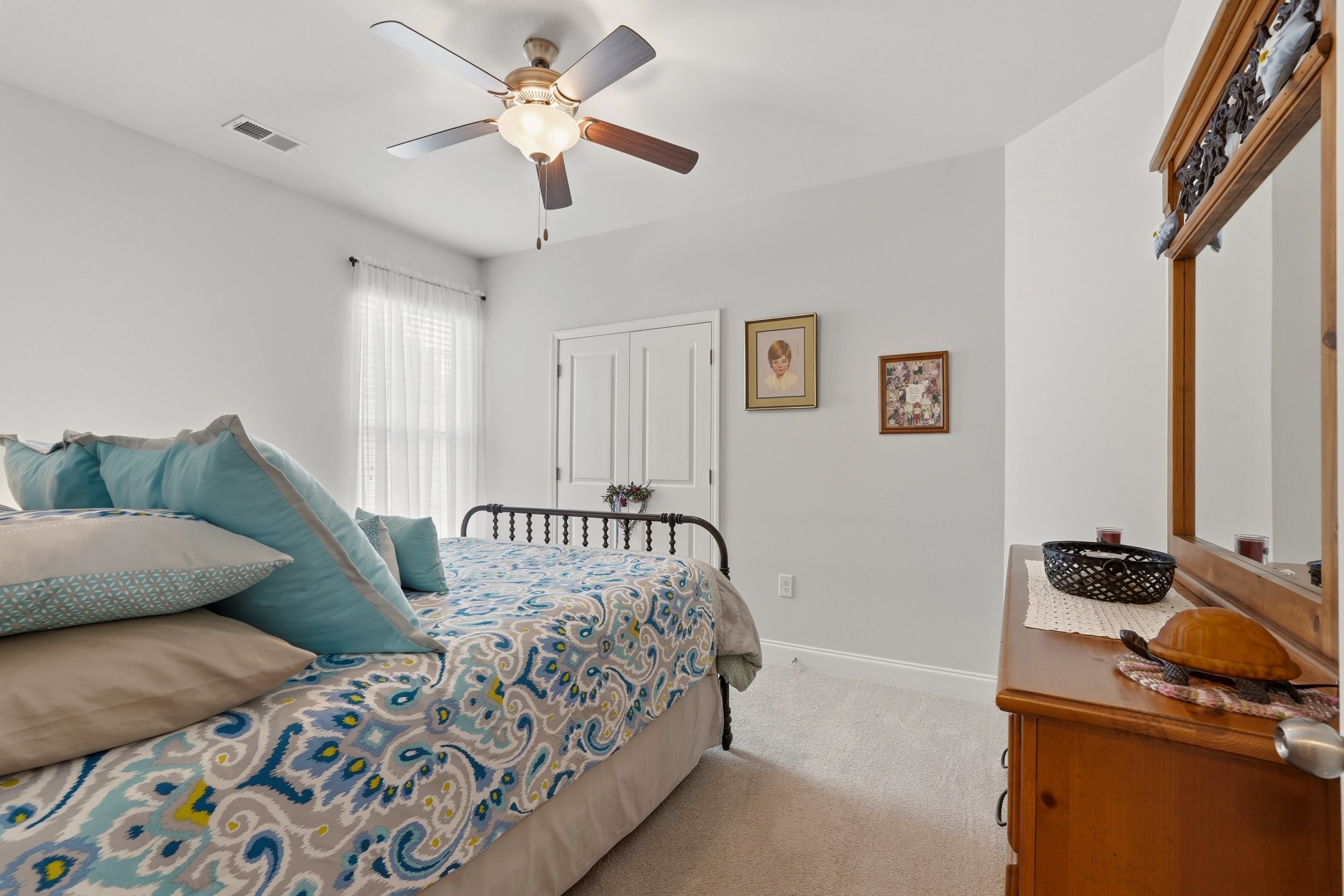 A bedroom with a bed covered in a patterned bedspread and multiple pillows, a wooden dresser with decorative items, framed pictures on the wall, a ceiling fan, and a window with sheer curtains.