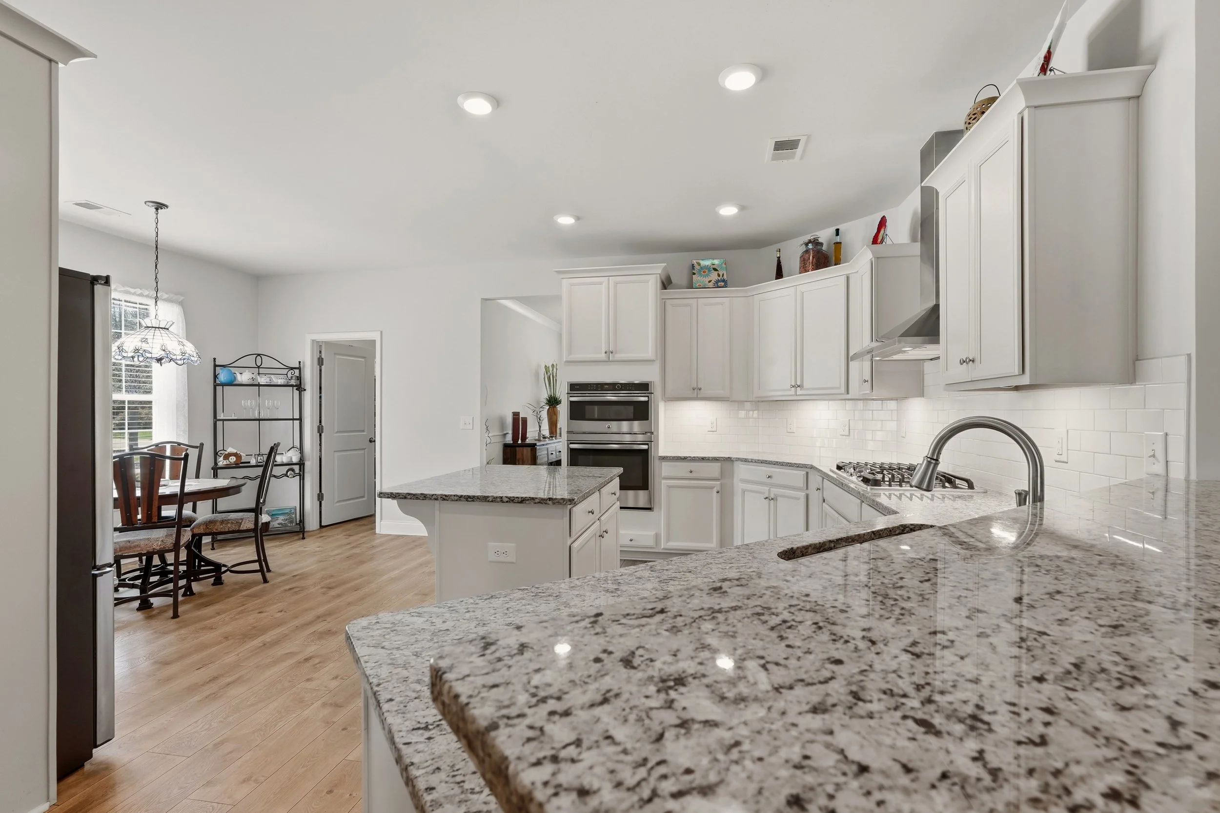 Open kitchen with granite countertops, white cabinets, stainless steel oven, and a dining area with wooden chairs and a black metal shelf. Recessed ceiling lights and a window with curtains are visible.