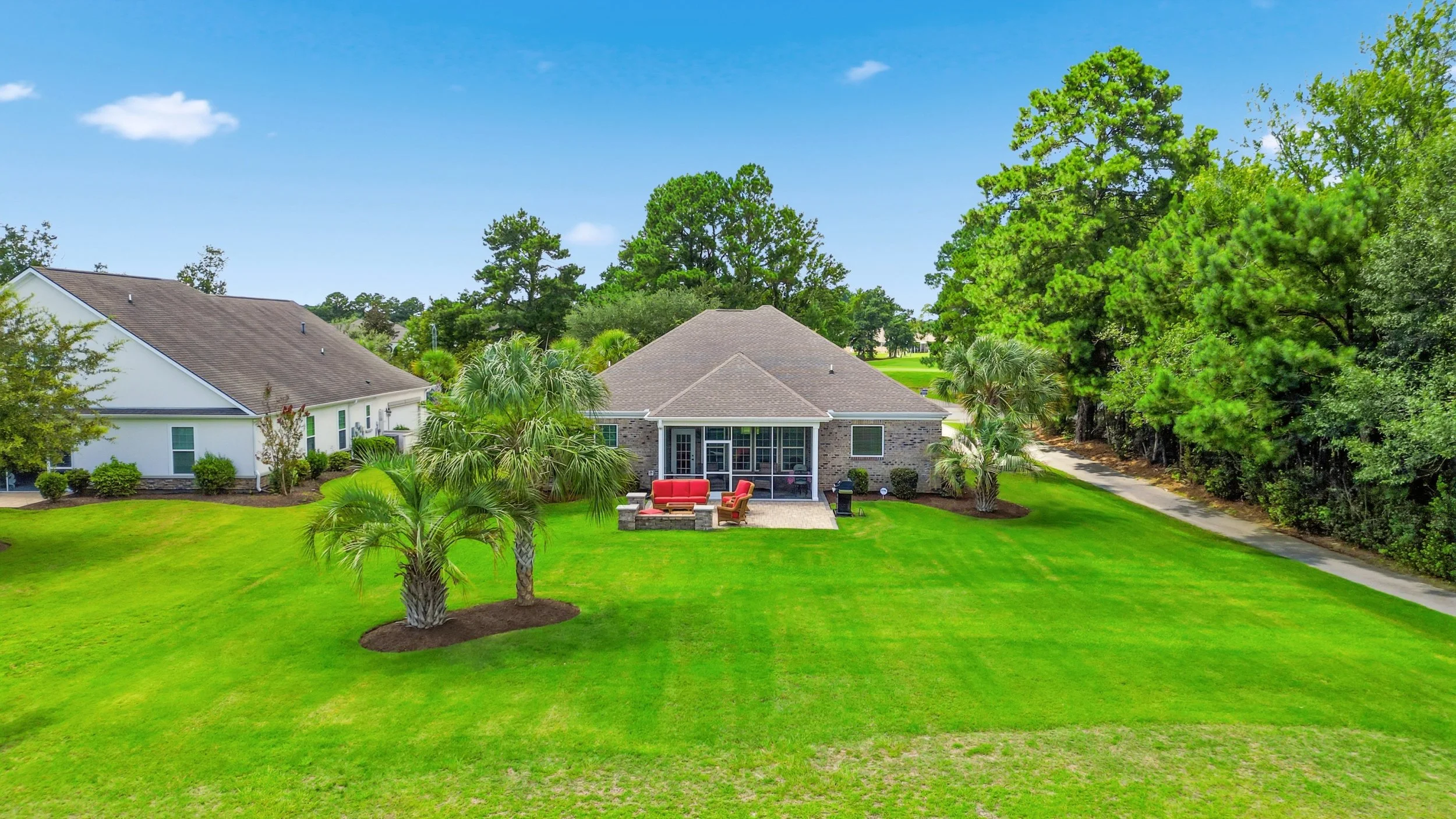 Backyard of a house with a well-maintained lawn, outdoor furniture, palm trees, and surrounded by tall green trees under a blue sky.