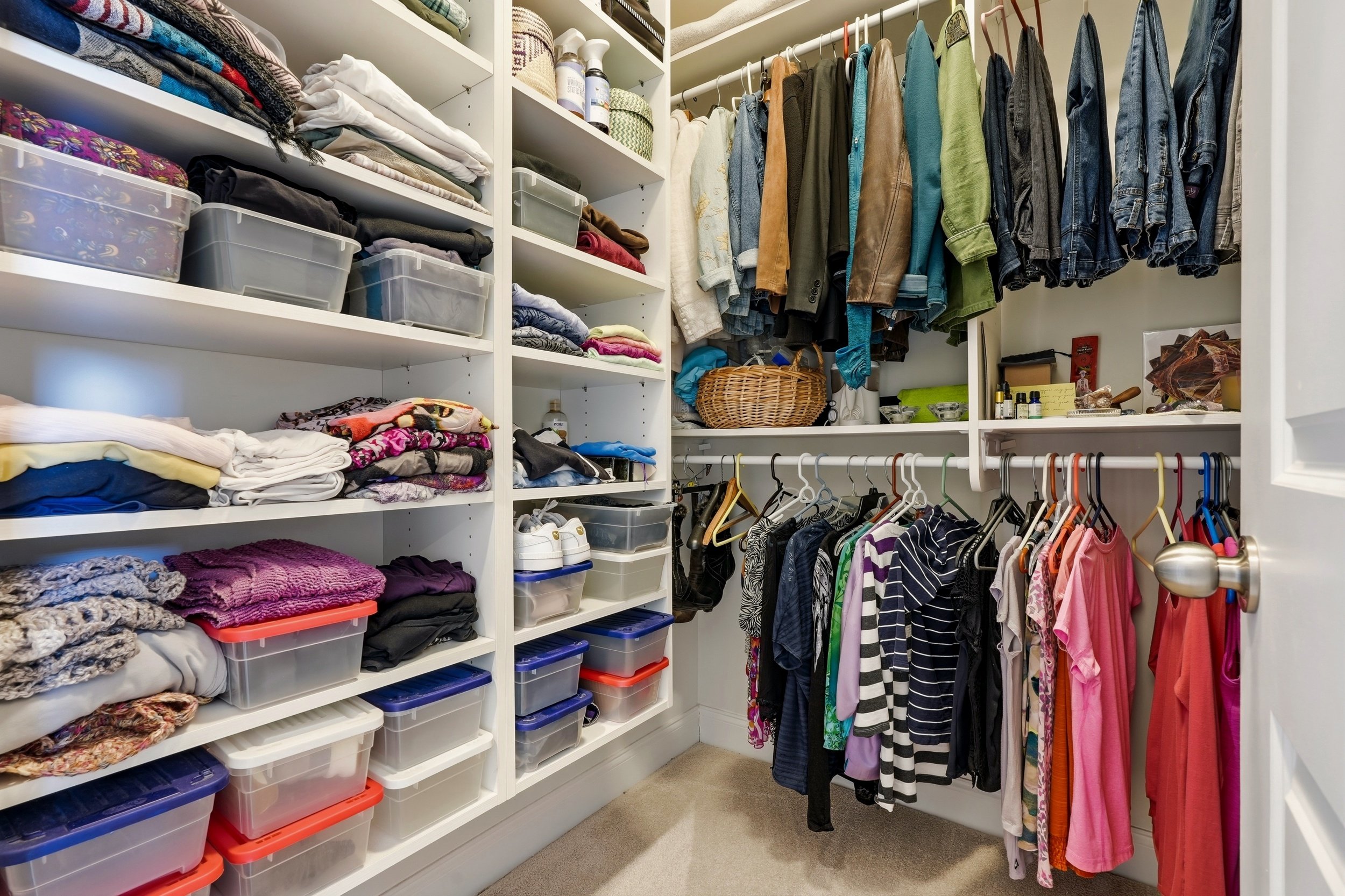A walk-in closet with white shelves filled with folded clothes, storage boxes, and accessories. Clothes hang on rods, including shirts, jackets, and dresses. The closet has beige carpeting and is well-organized.