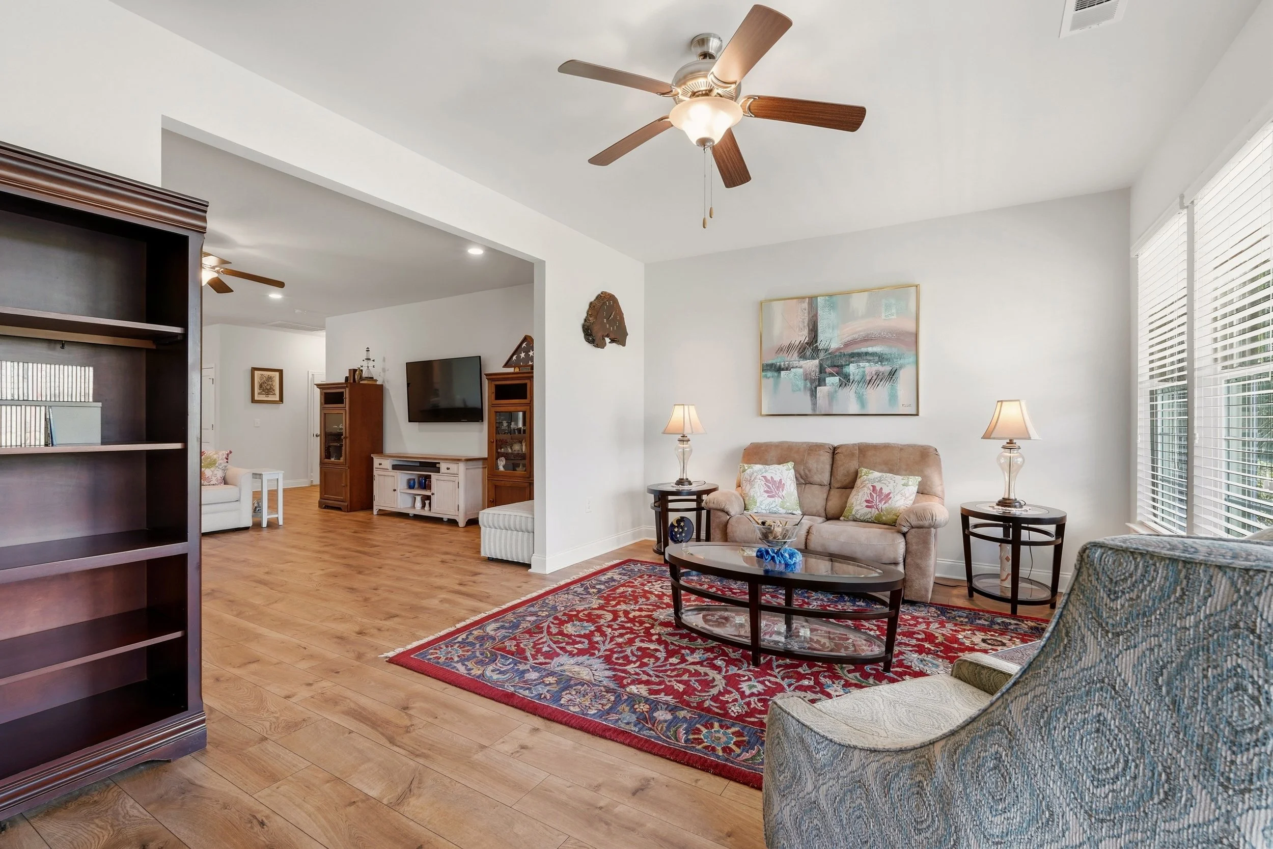 Living room with hardwood floor, beige sofa, armchair, glass coffee table, bookshelf, wall art, TV, and ceiling fan.