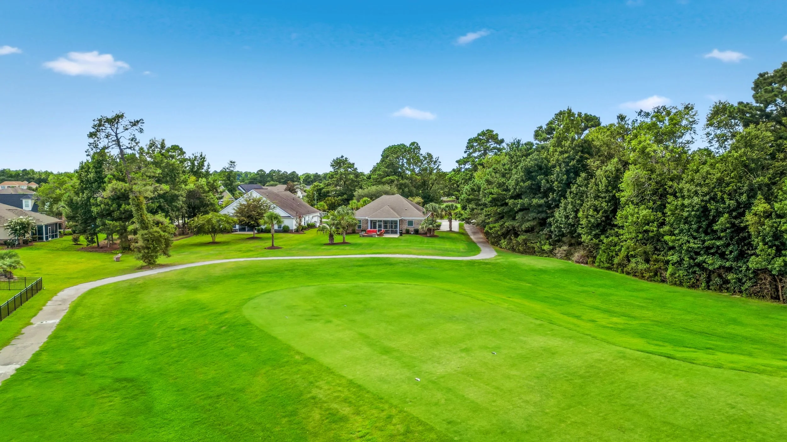A lush green golf course with a curved pathway, surrounded by trees and residential houses under a bright blue sky.