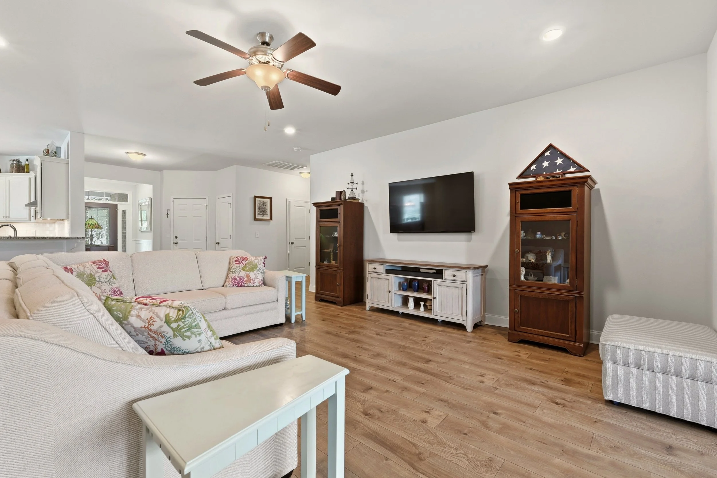 Living room with white sofa, colorful pillows, wooden floor, wall-mounted TV, and wooden cabinets with patriotic display