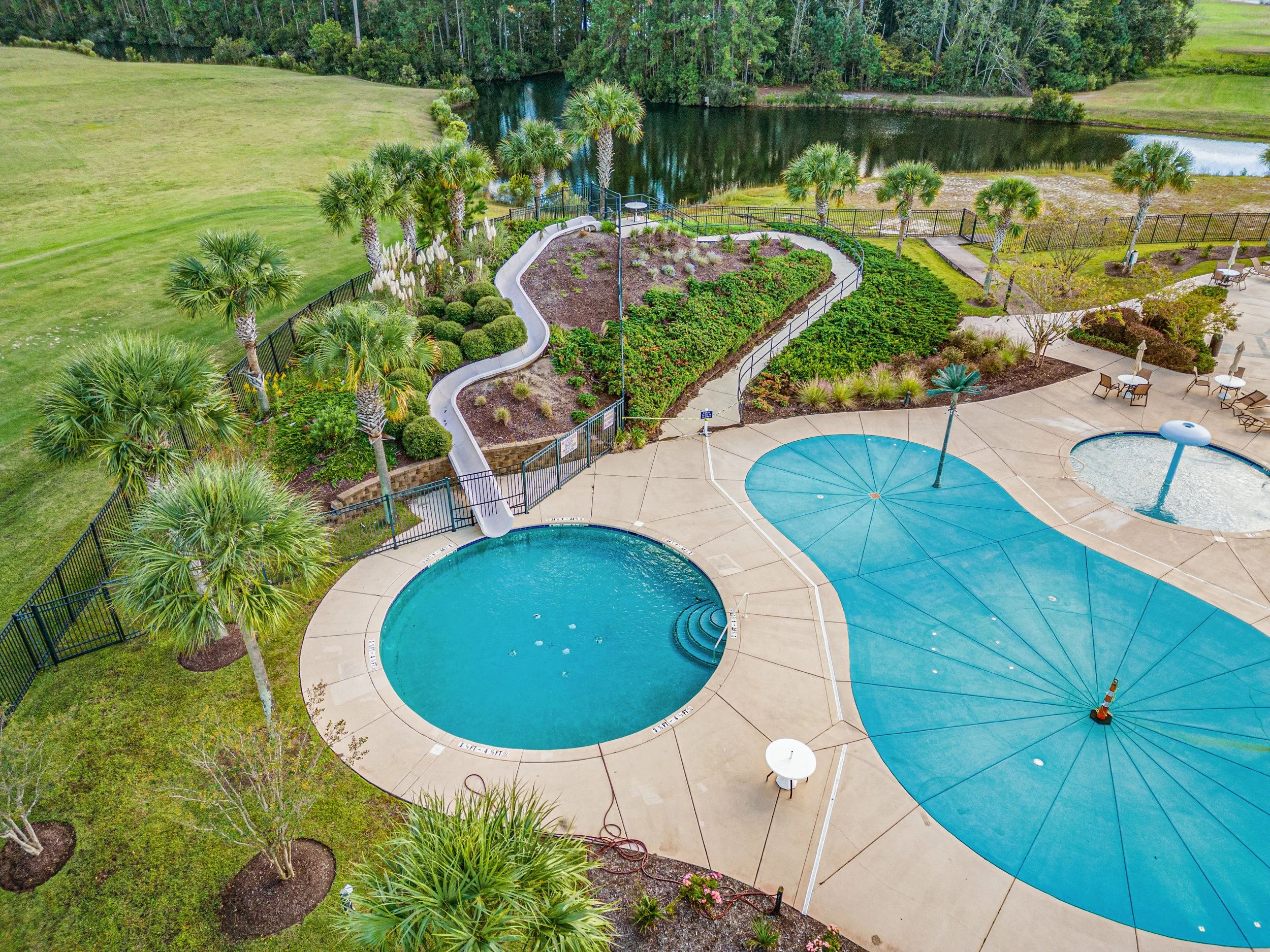 An outdoor swimming pool area with a large pool, a smaller hot tub, and a blue shaded umbrella. Surrounding the area are trees, palm trees, and a landscaped garden with pathways and seating.