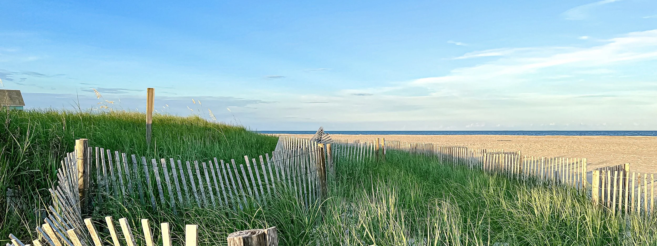 A beach scene with green grassy dunes, a sandy shoreline, and a blue ocean under a partly cloudy sky. Wooden fence posts line the dunes, leading to the beach.