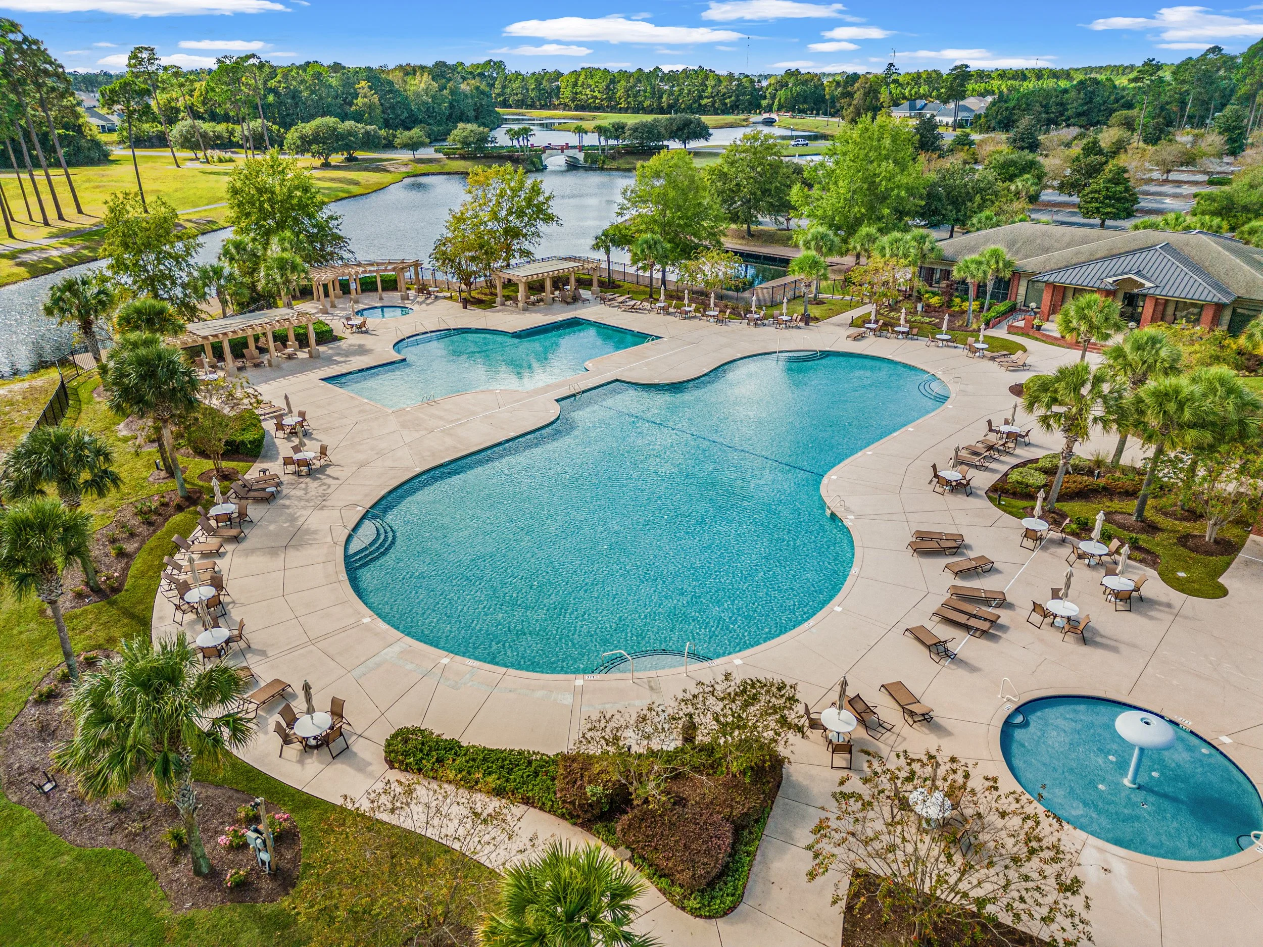 Aerial view of an outdoor swimming pool area with lounge chairs, umbrellas, and lush green trees, surrounded by a concrete patio, overlooking a water body and a green landscape.