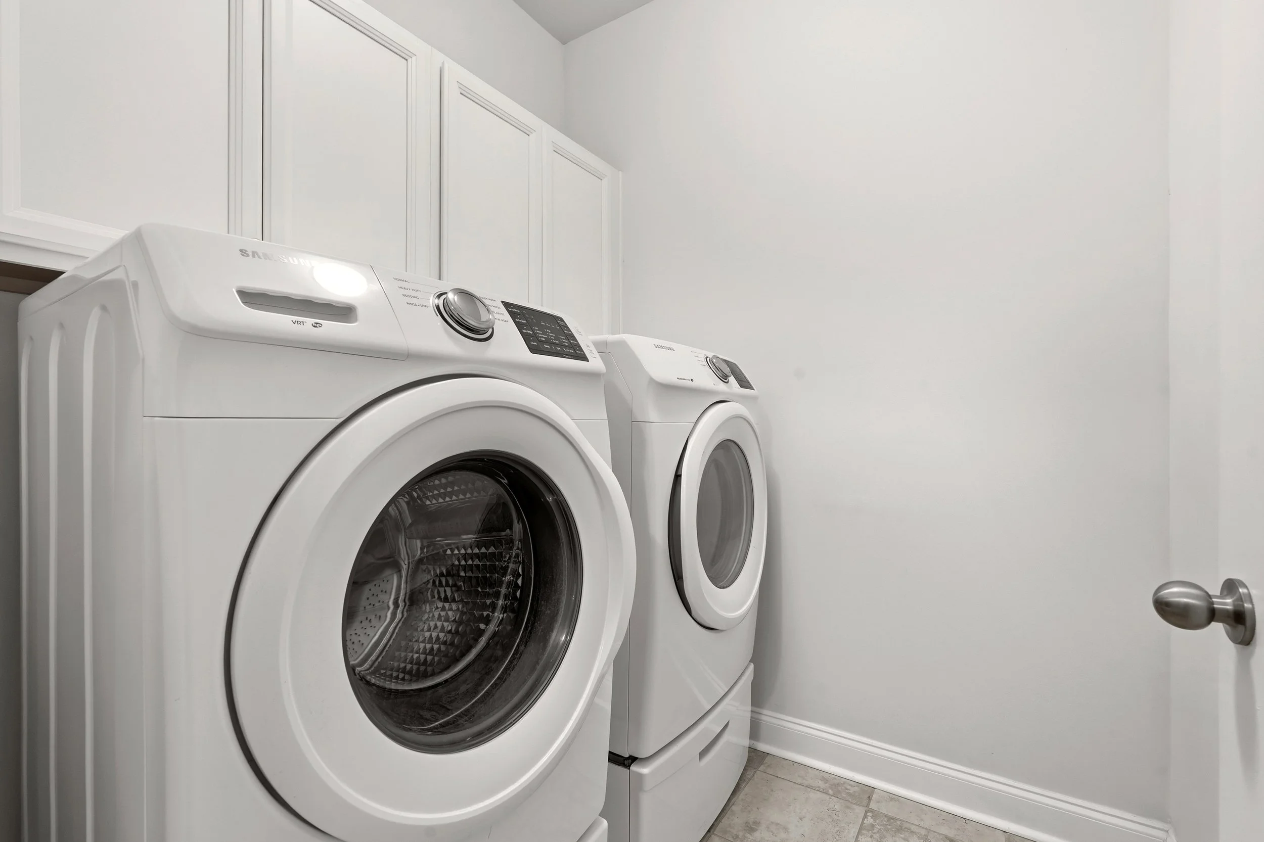 A side view of a laundry room with a white washing machine and dryer placed side by side against a white wall, with cabinet storage above them and a door on the right.