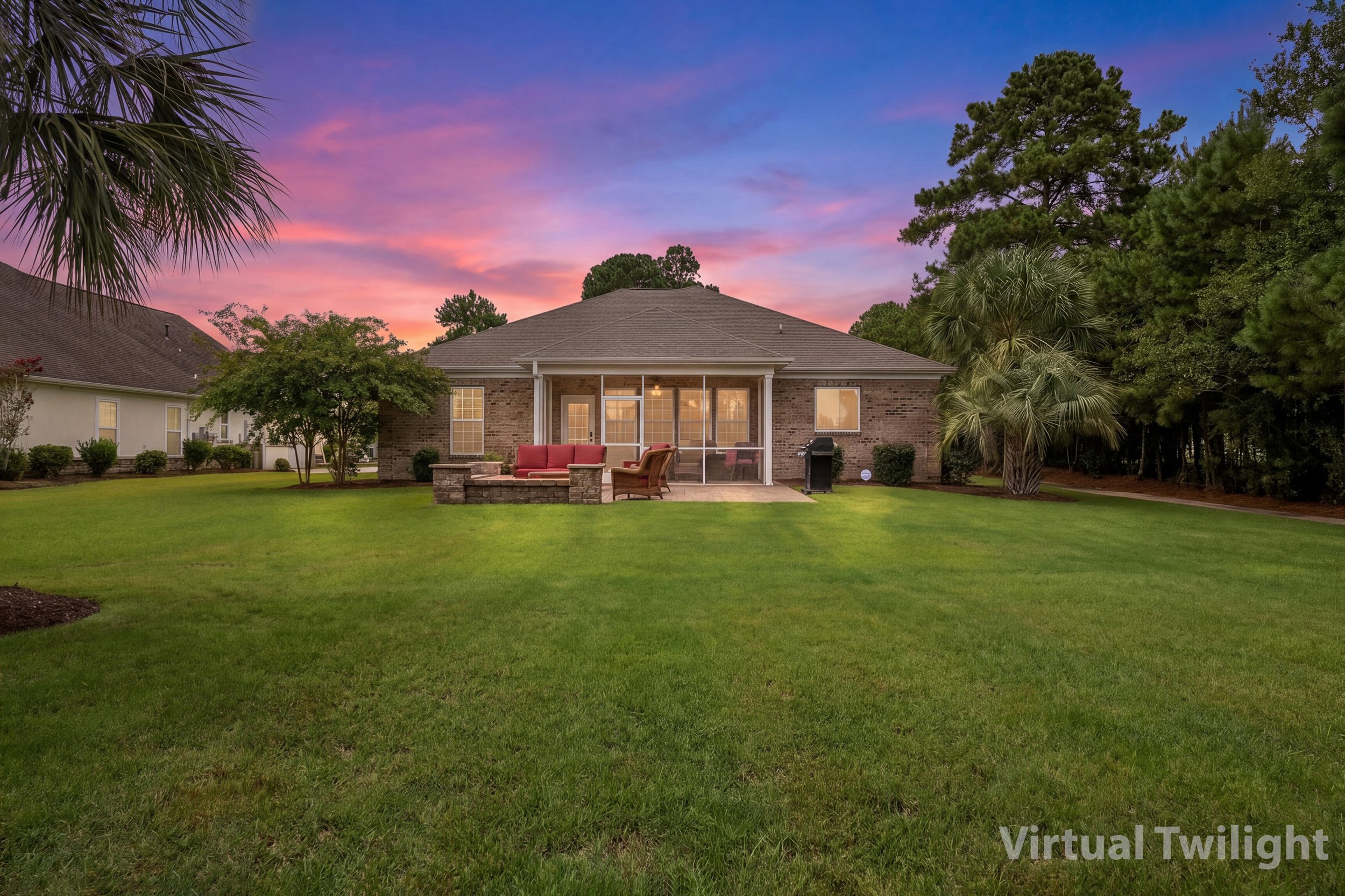 A backyard view during sunset featuring a grassy lawn, a brick house with a screened porch, outdoor furniture, trees, and a colorful sky with pink and purple clouds.