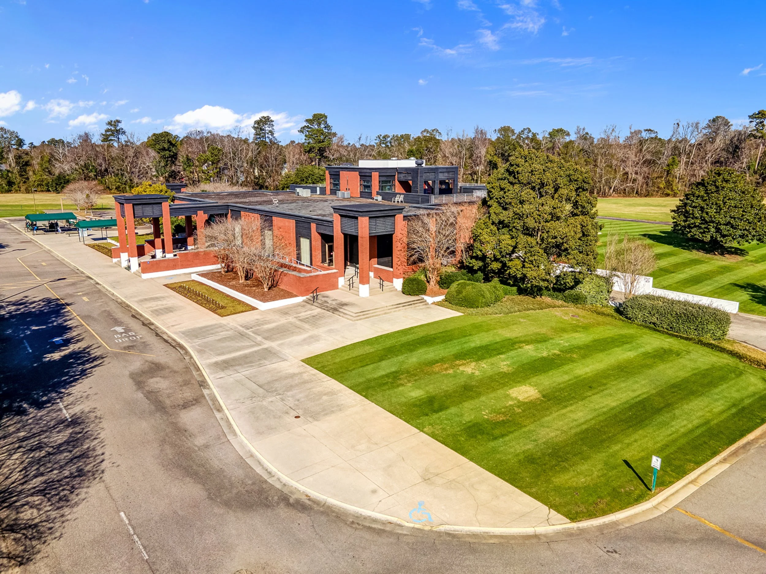 An aerial view of a modern brick building surrounded by trees, a parking lot, and grassy areas, under a bright blue sky with scattered clouds.