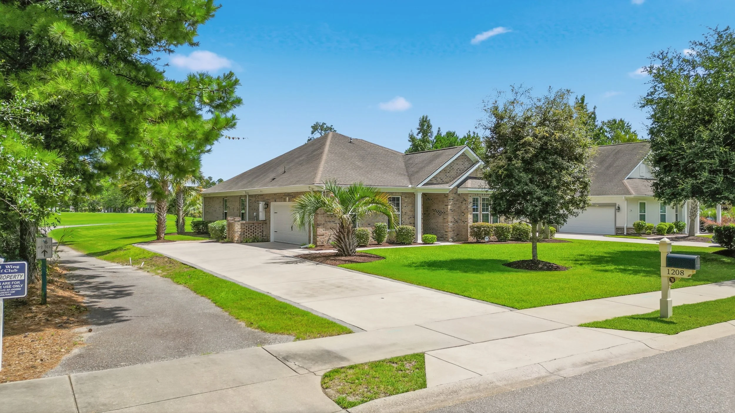 A suburban house with a brick exterior, a garage door, and a well-maintained lawn. There are trees and shrubs around the property, clear skies, and a sidewalk in front.