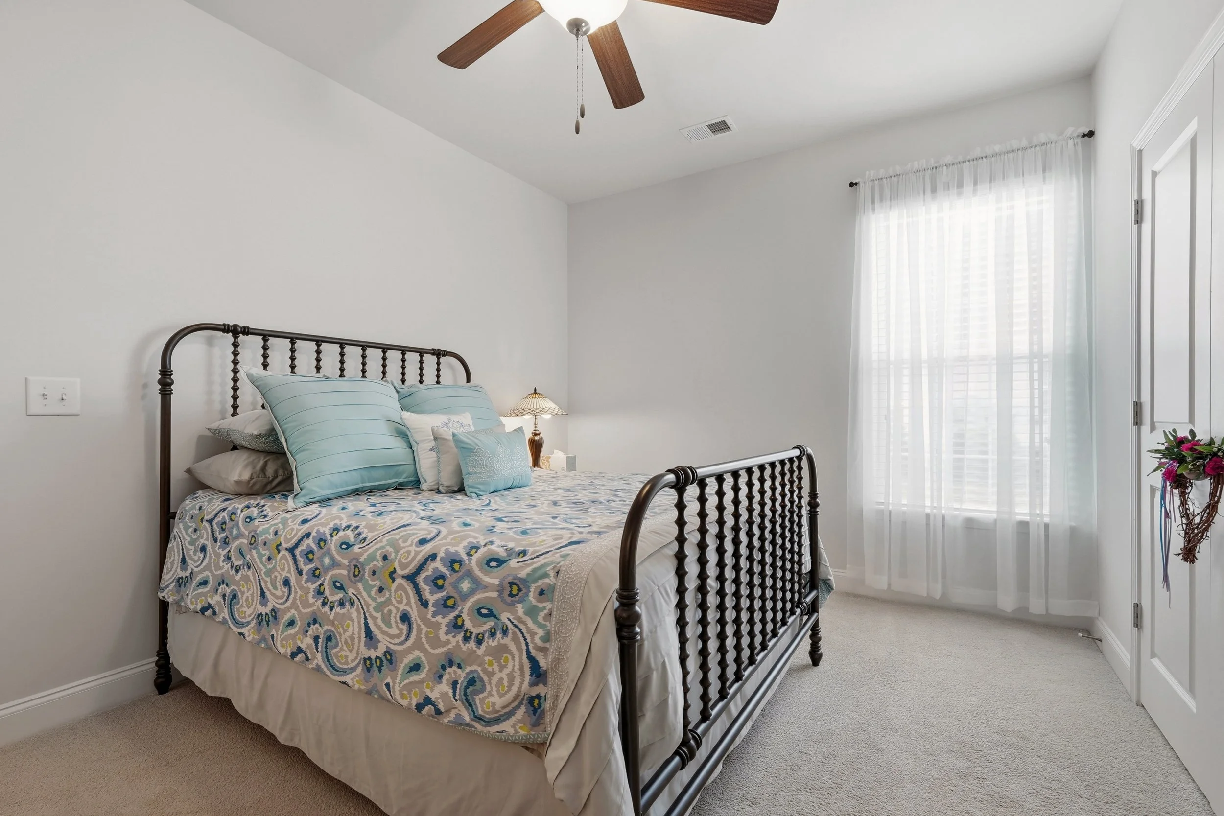 A bedroom featuring a metal bed frame with decorative finials, adorned with blue and patterned pillows, a bedside lamp with a stained glass-style shade, white walls, a window with sheer white curtains, ceiling fan with wooden blades, and light-colore