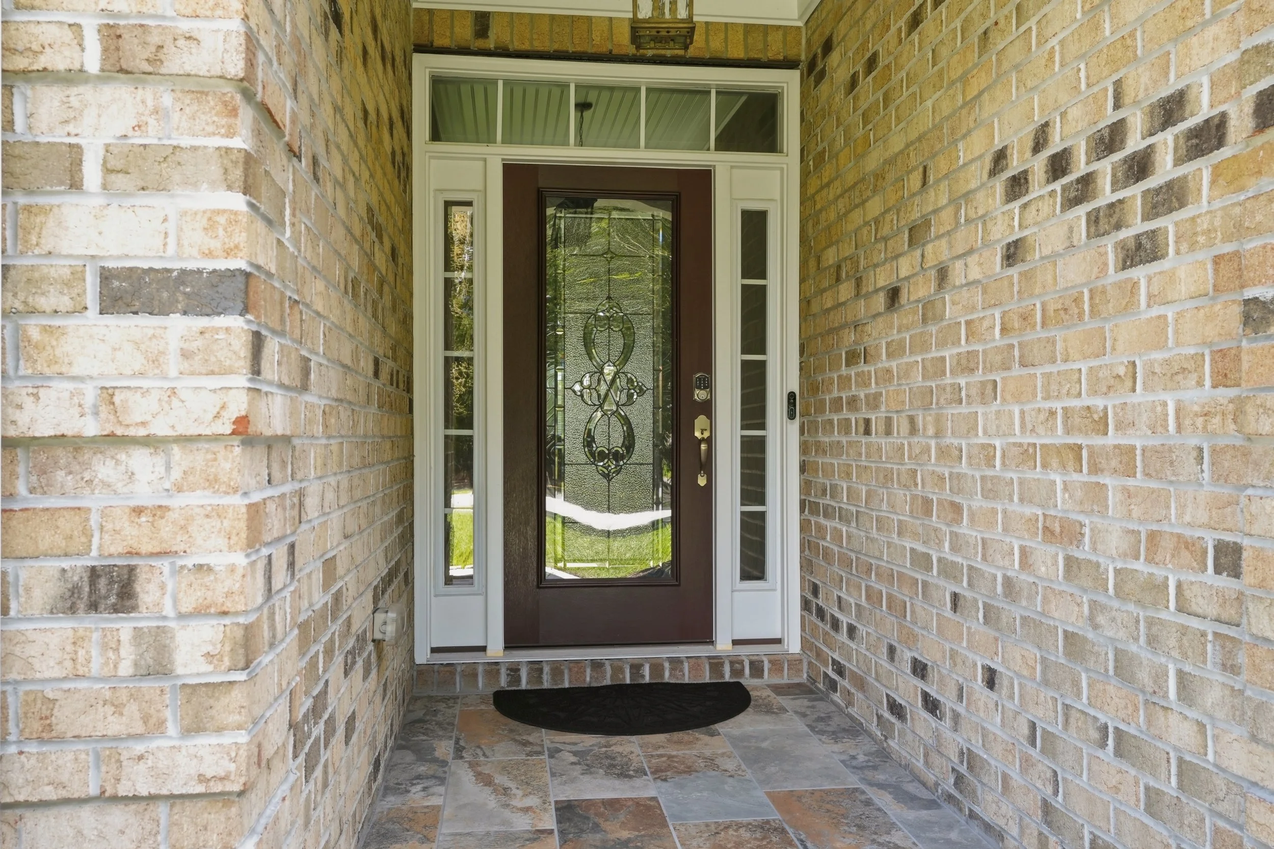 Front porch with brick walls, a front door with decorative glass, sidelights, and a transom window, with tiled flooring and a black doormat.