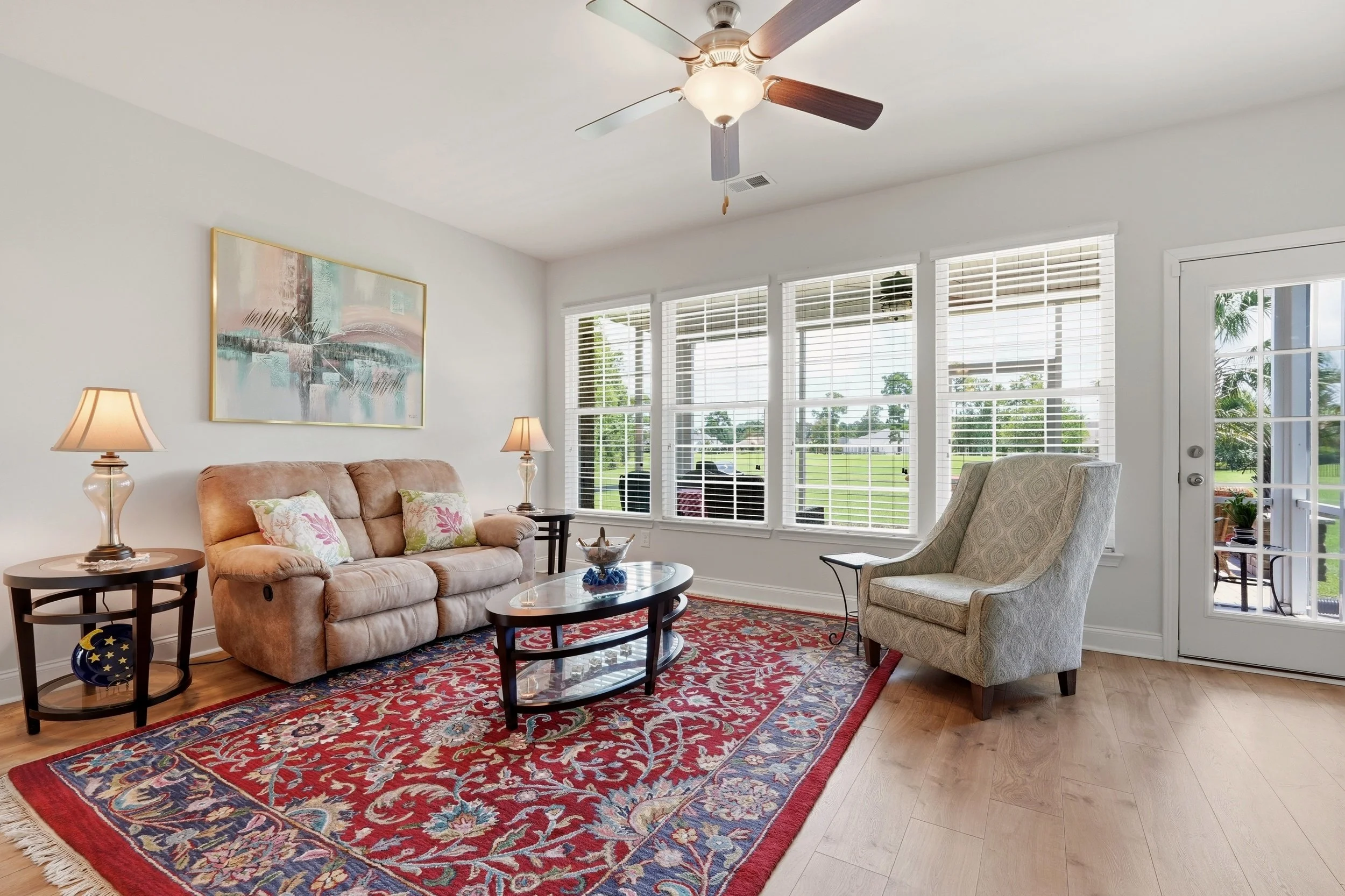 Living room with beige sofa, two side tables with lamps, an armchair, a glass coffee table, a patterned red area rug, large windows with white blinds, a ceiling fan, and a wall art painting.
