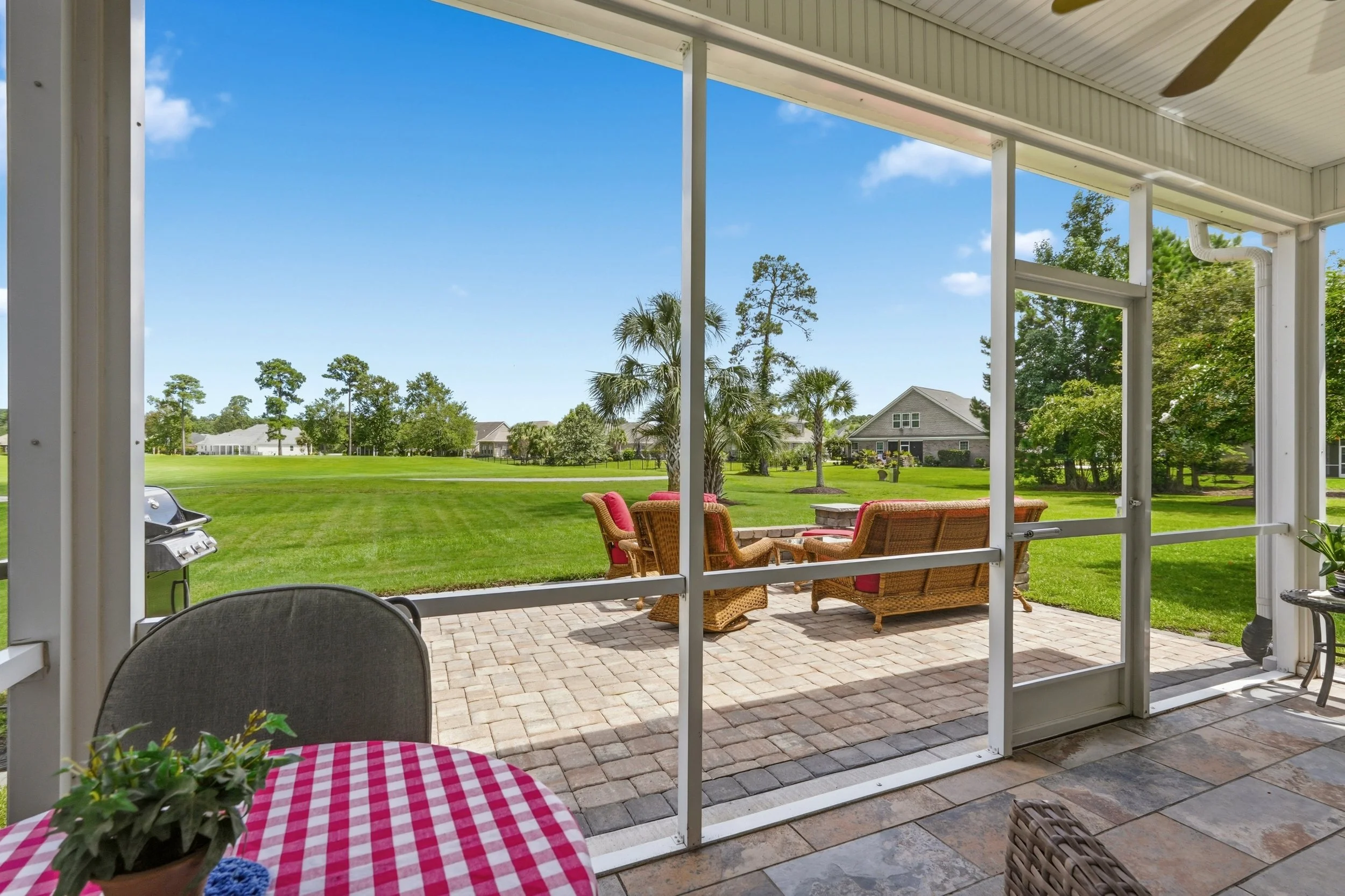 View from a house porch showing a backyard with green grass, golf course view, trees, and neighboring houses under a blue sky with a few clouds.