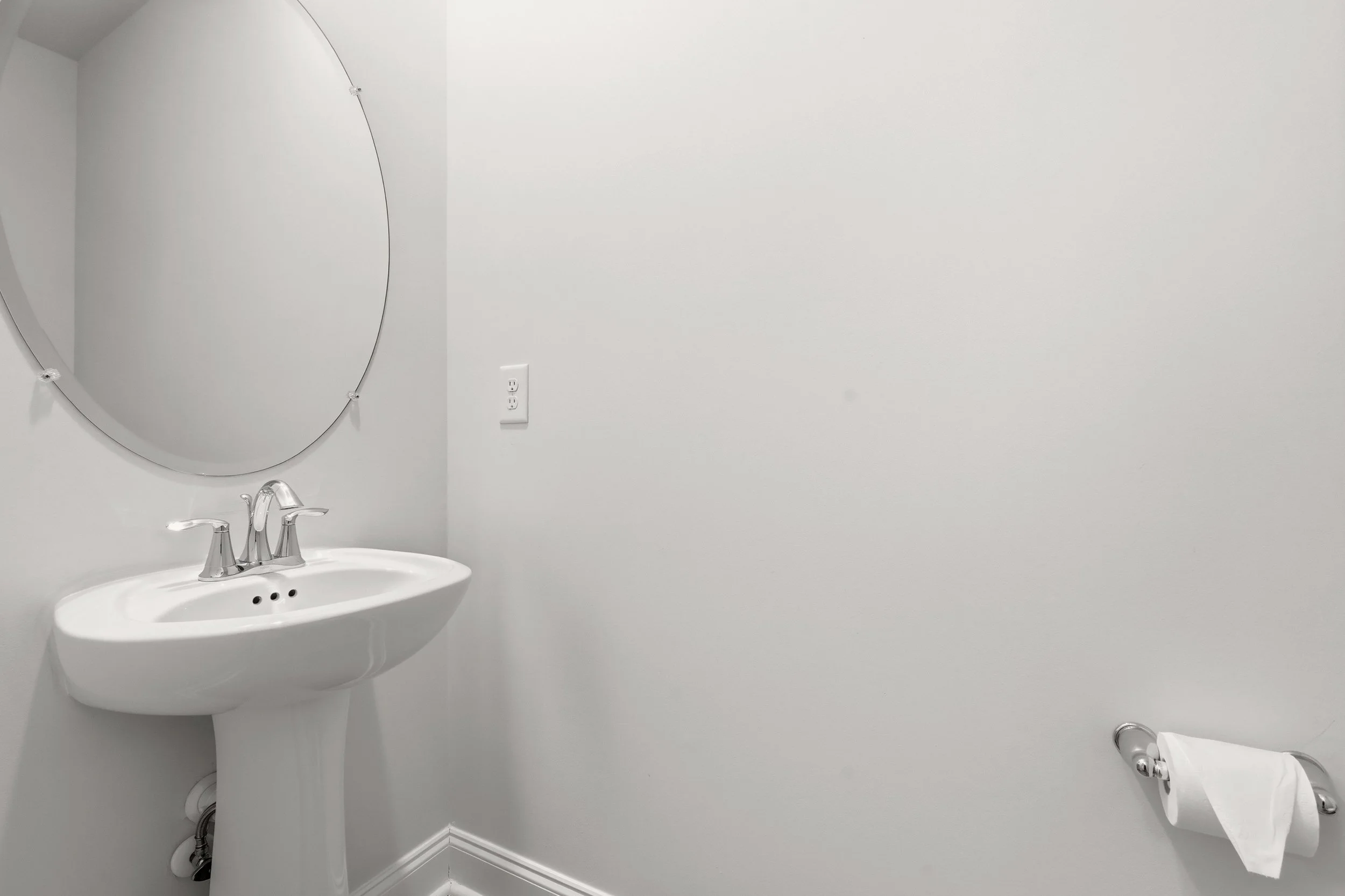 Bathroom with white sink, oval mirror, white walls, electrical outlet, and toilet paper roll.