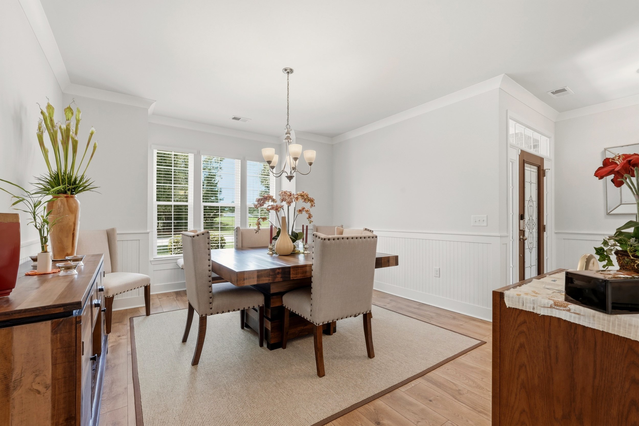 A dining room with a wooden table surrounded by beige upholstered chairs, decorated with pink flowers in a beige vase, a sideboard with plant and decorative items, large windows with blinds, and a chandelier hanging from the ceiling.