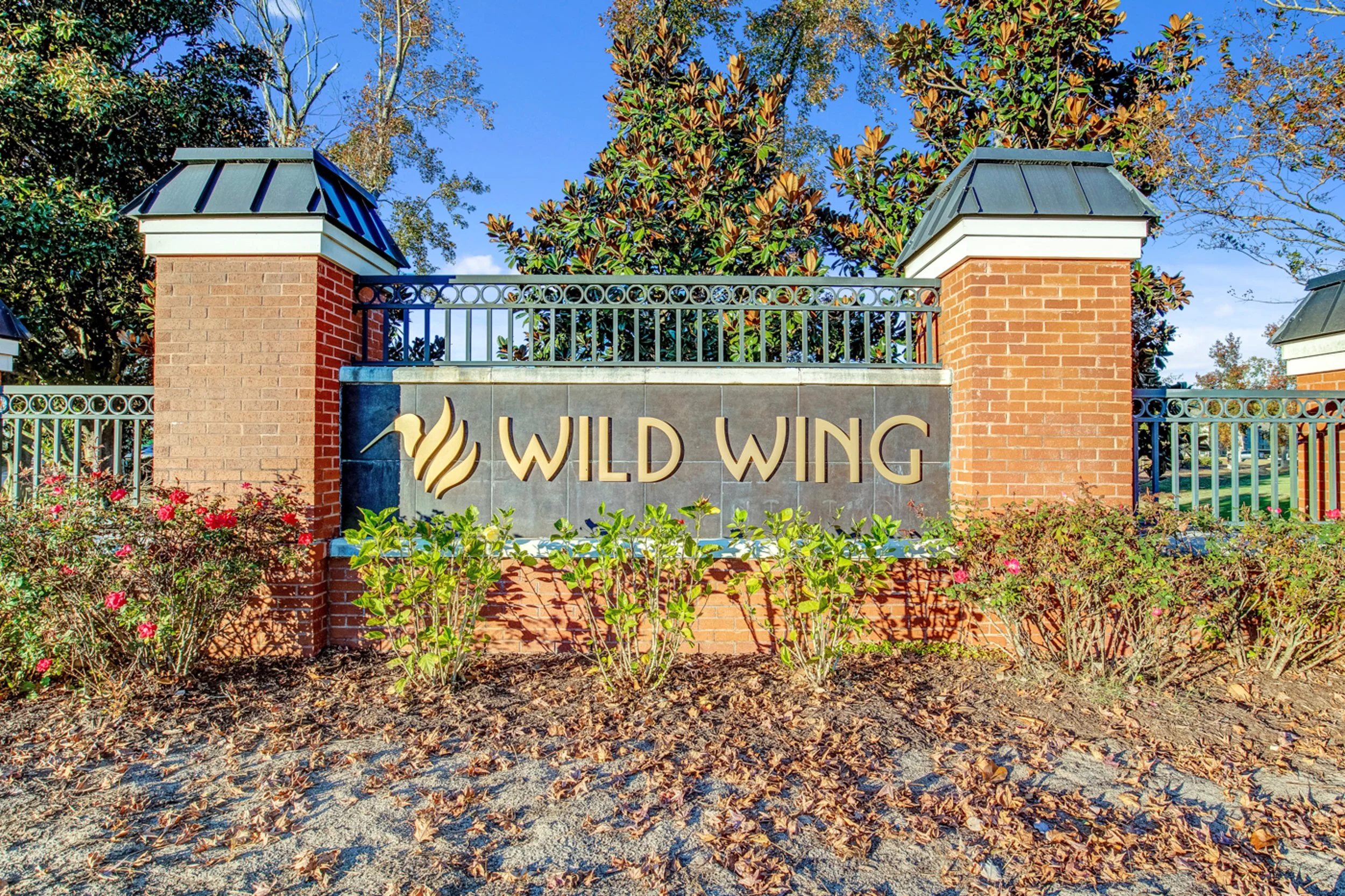 Image of a gated entry with a sign that reads "Wild Wing" in front of brick pillars, with trees and bushes in the background, under a clear blue sky.