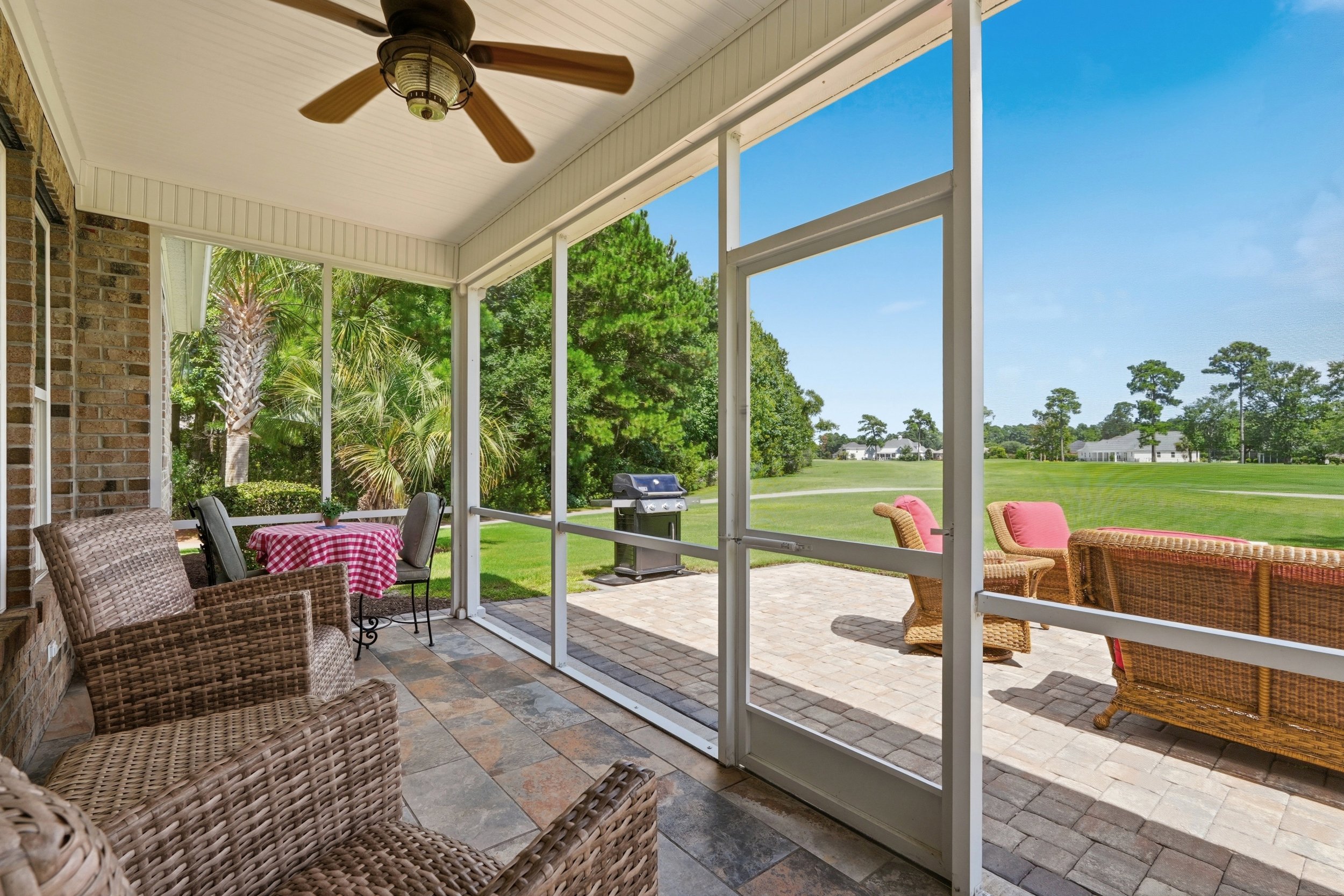 Covered patio with wicker chairs and a small round table with a red-and-white checkered tablecloth, overlooking a green backyard with golf course view, trees and a grill.
