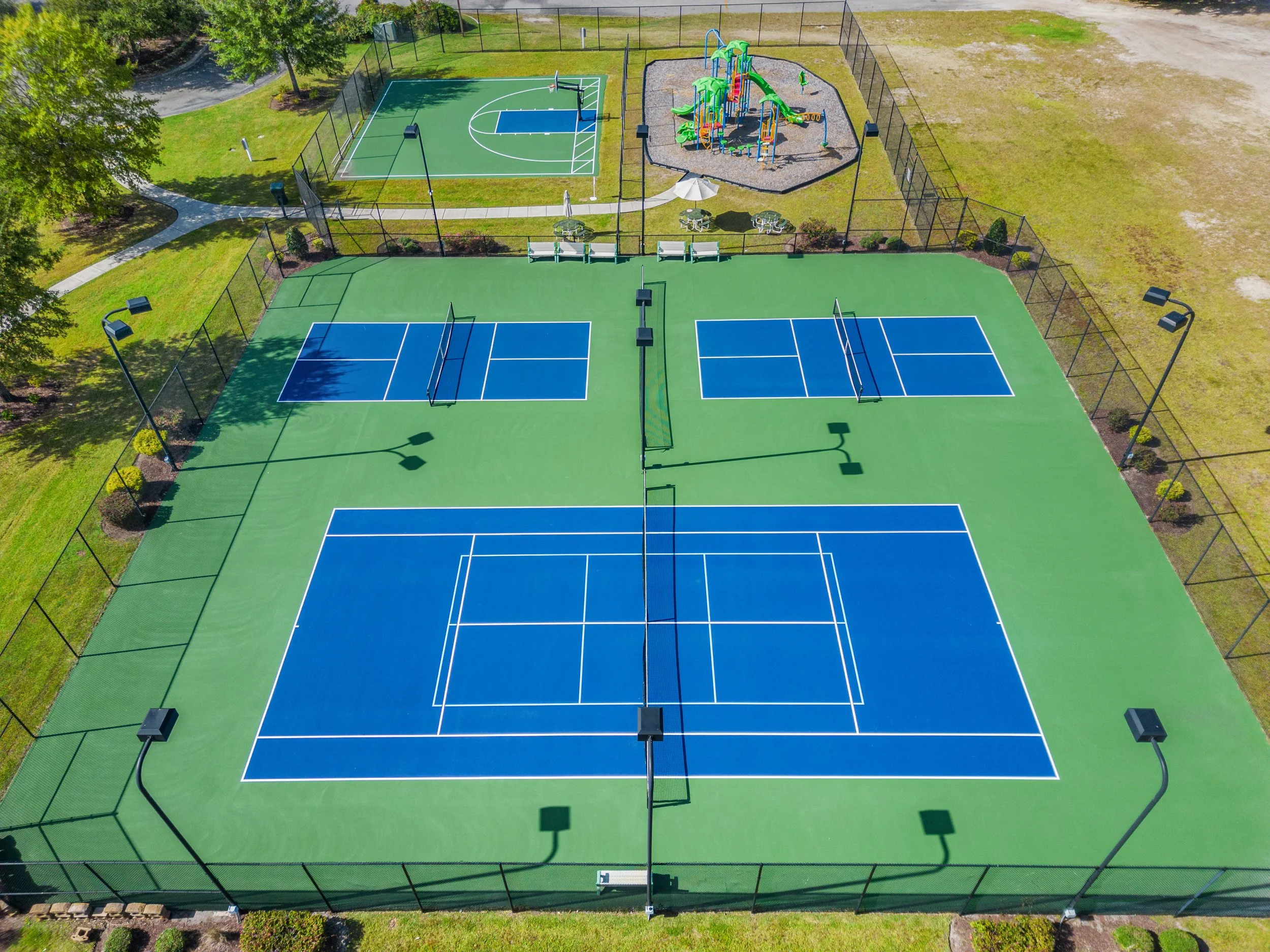 Aerial view of a sports complex featuring two tennis courts with blue playing surfaces and a basketball court, a children's playground with slides and climbing structures, surrounded by green grass, trees, and pathways.