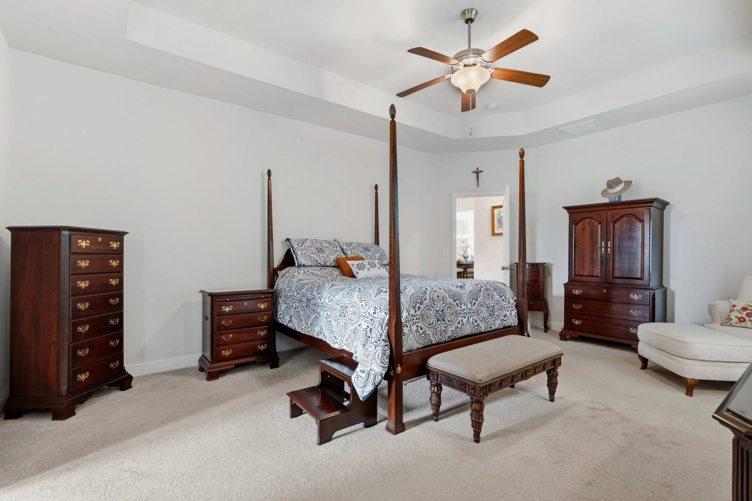 A bedroom featuring a four-poster bed with patterned bedding, wooden furniture including dressers and a wardrobe, a ceiling fan with wooden blades, a crucifix on the wall, and a hat on top of the wardrobe.