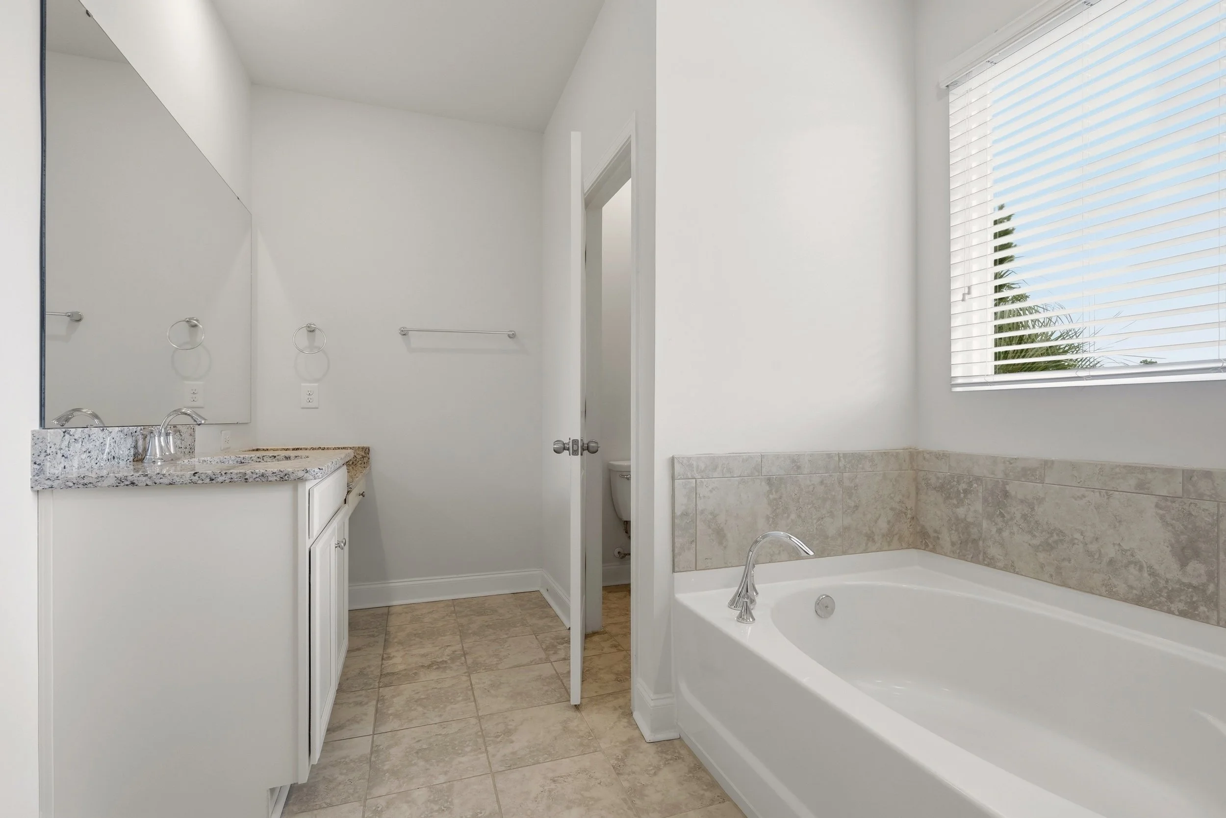 A bathroom with a white vanity, granite countertop, mirror, towel rings, towel bar, toilet, bathtub with tiled surround, and a window with blinds letting in natural light.
