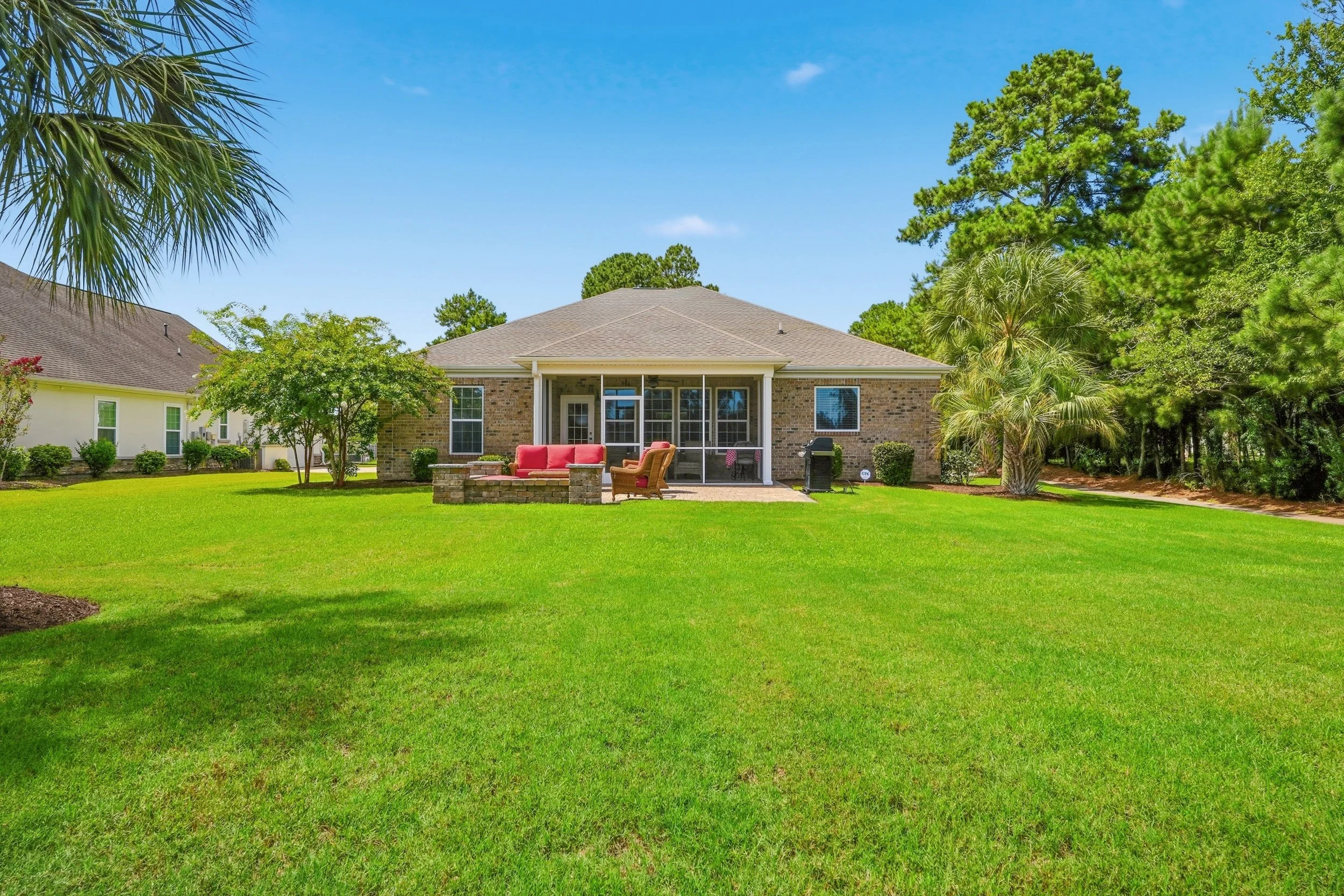 Backyard with green grass, trees, and a single-story brick house with a patio and outdoor furniture.