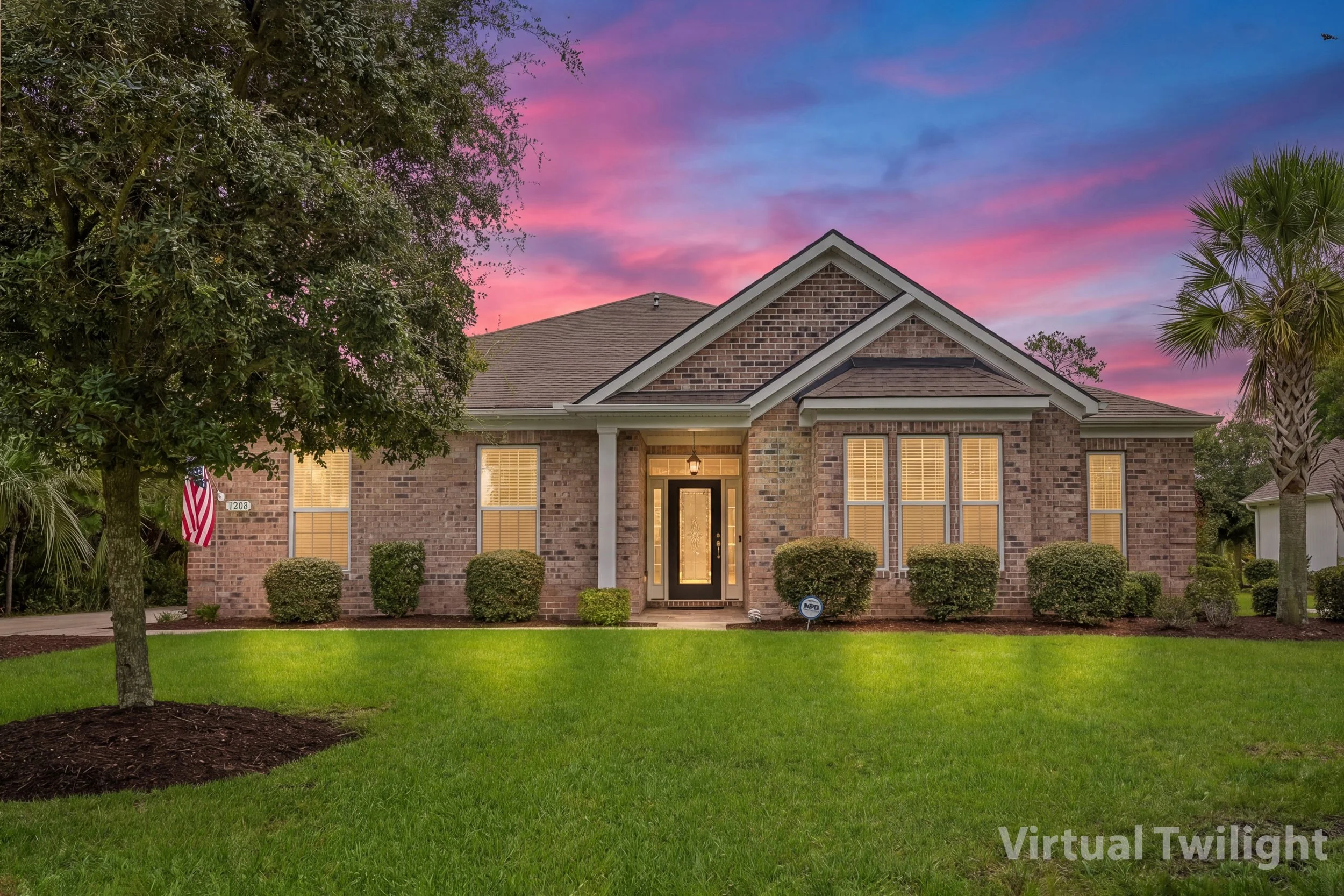 A brick house with front windows and a front door under a gabled roof, surrounded by a well-maintained lawn, trees, and shrubs, during sunset with colorful pink and purple sky.