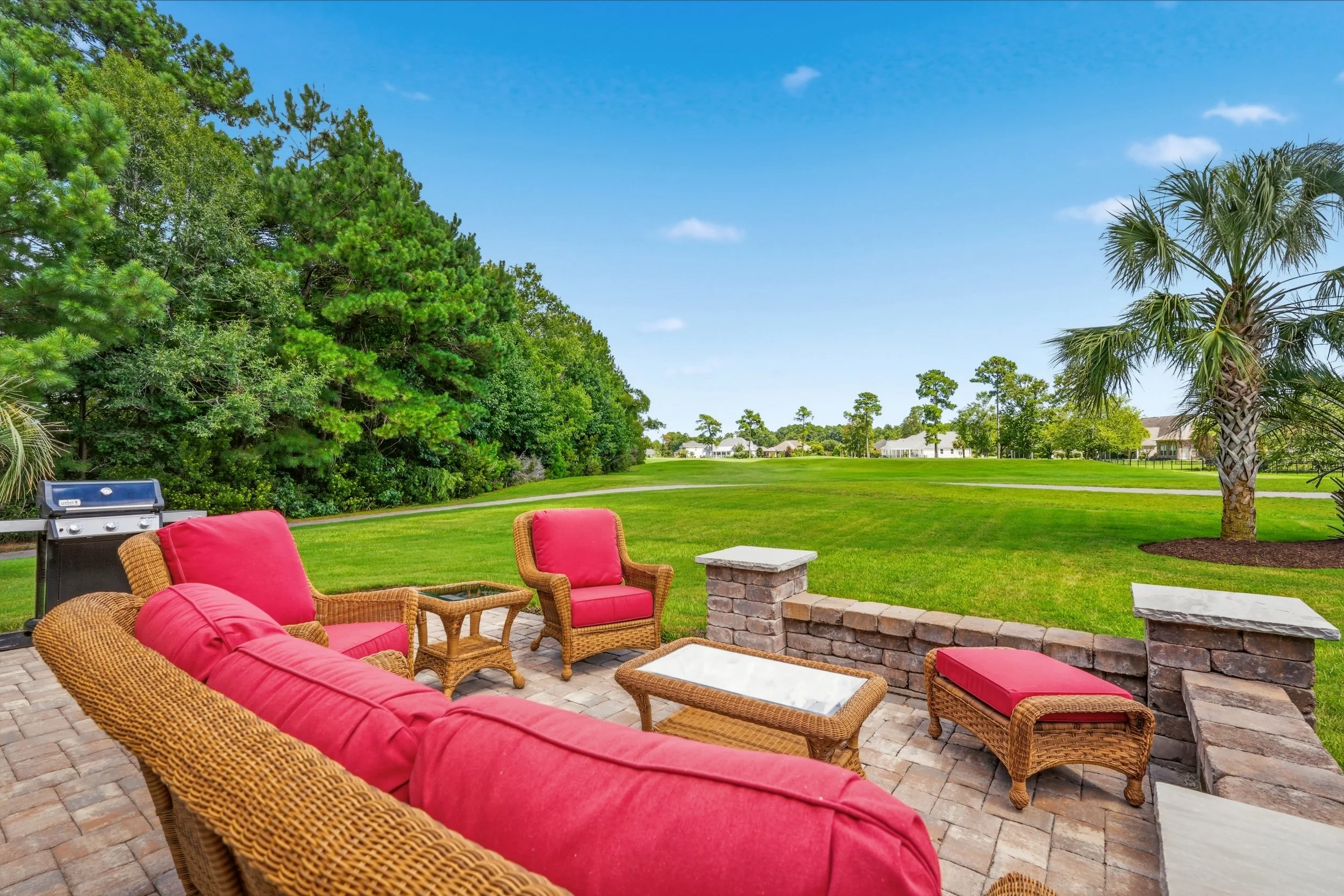 Backyard patio with red cushioned wicker furniture, a barbecue grill, a stone fire pit, golf course view, and a lush green lawn with trees and houses in the background under a blue sky with some clouds.