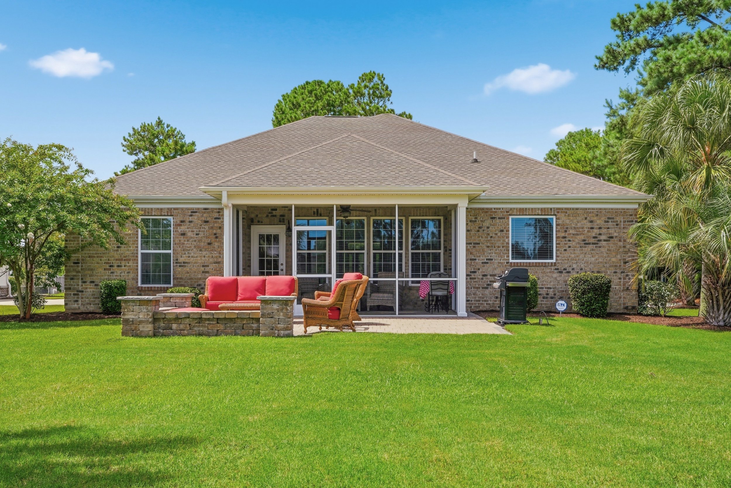Backyard view of a brick house with a covered patio, patio furniture, a grill, and a green lawn surrounded by trees under a blue sky with clouds.