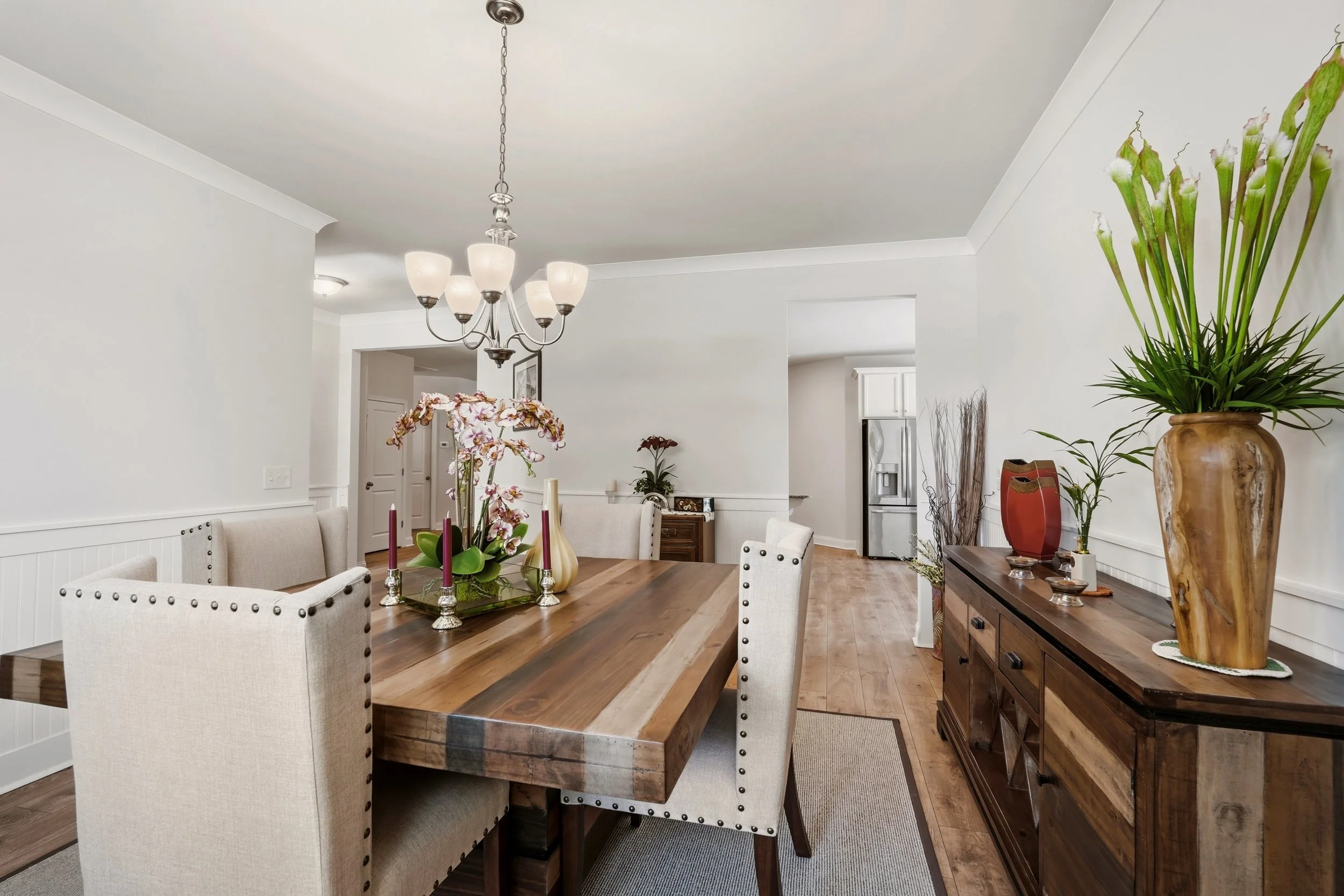 Dining room with a wooden table, beige upholstered chairs, floral centerpiece, large plants in pots, chandelier, and sideboard against white walls.