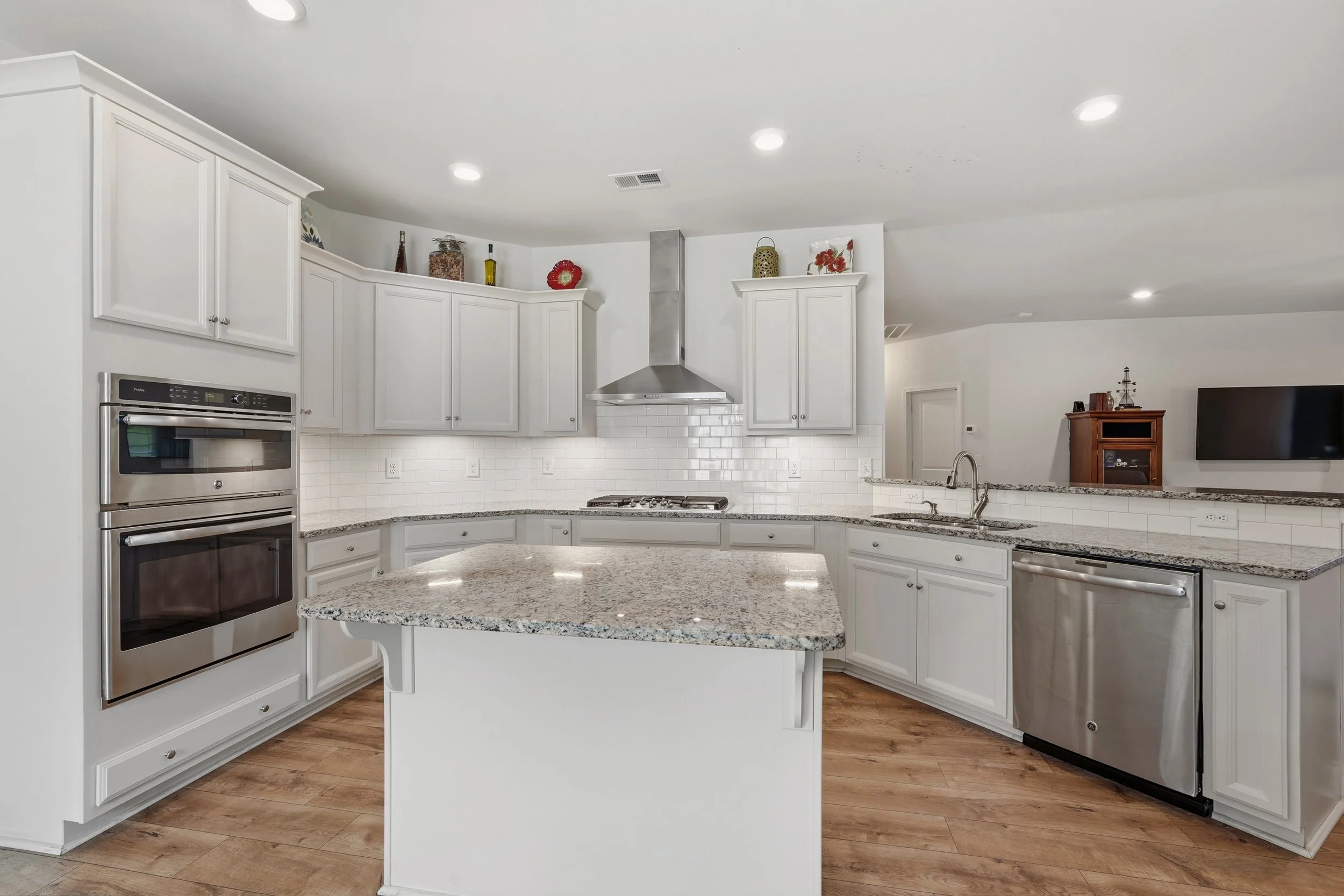 Modern white kitchen with granite countertops, stainless steel appliances, and decorative items on upper cabinets, overlooking a living area with a TV and wooden cabinet.