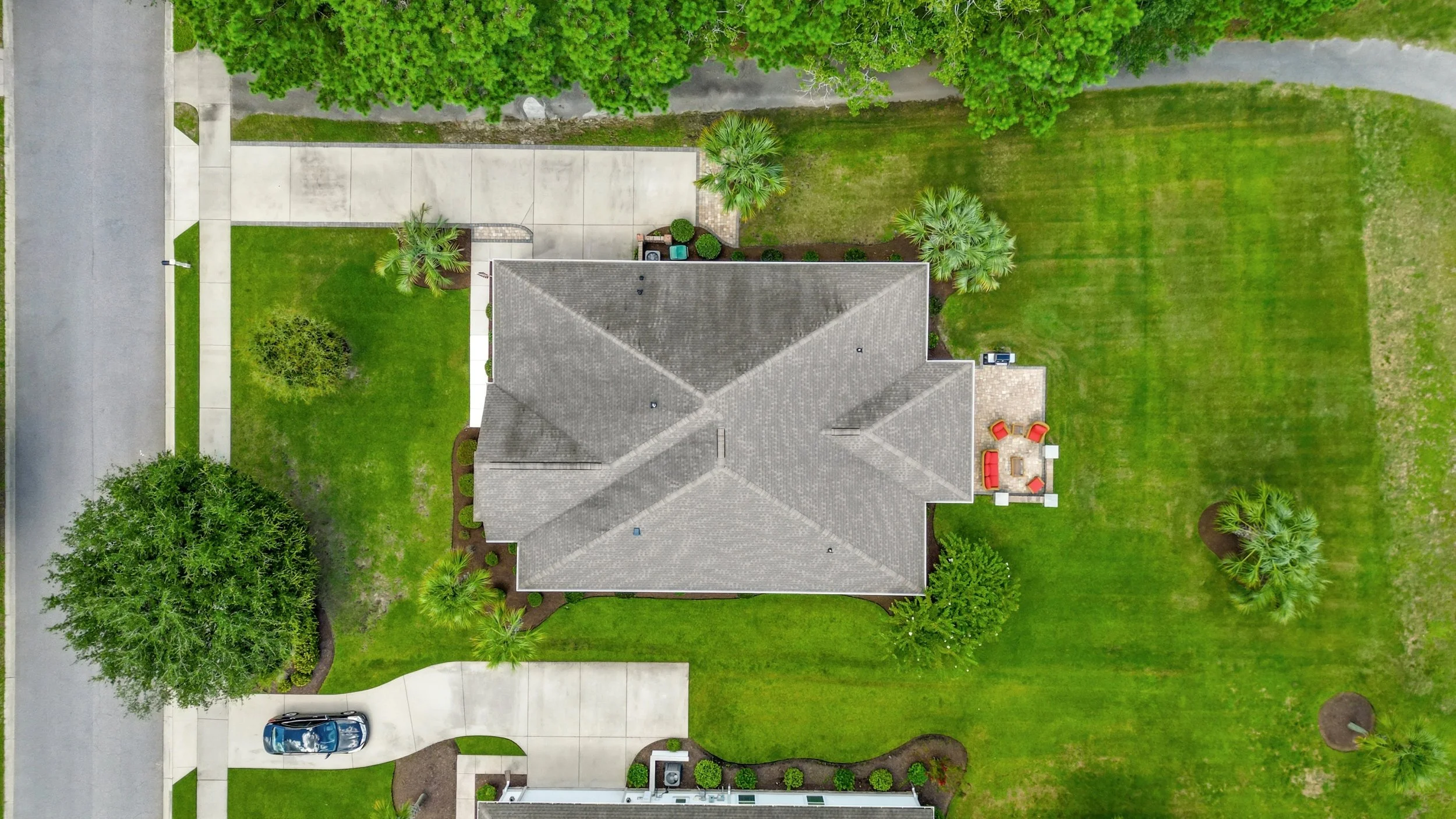 An aerial view of a house with a gray roof, surrounded by green lawn, trees, and a concrete driveway with a blue car parked near the door. There is a small backyard patio with red chairs and a table.