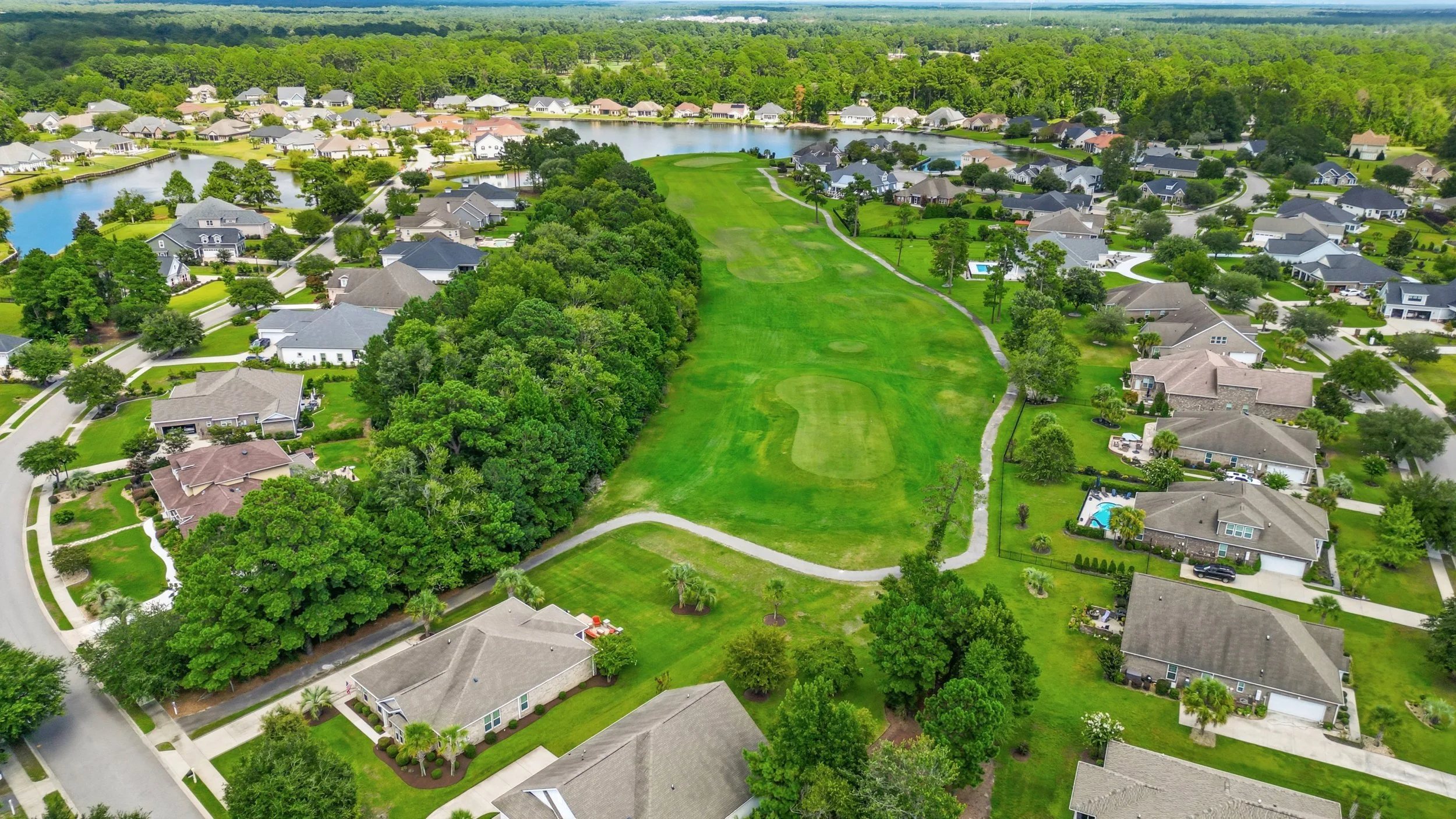 An aerial view of a suburban neighborhood with houses and yards, a golf course, a lake, and lush greenery.