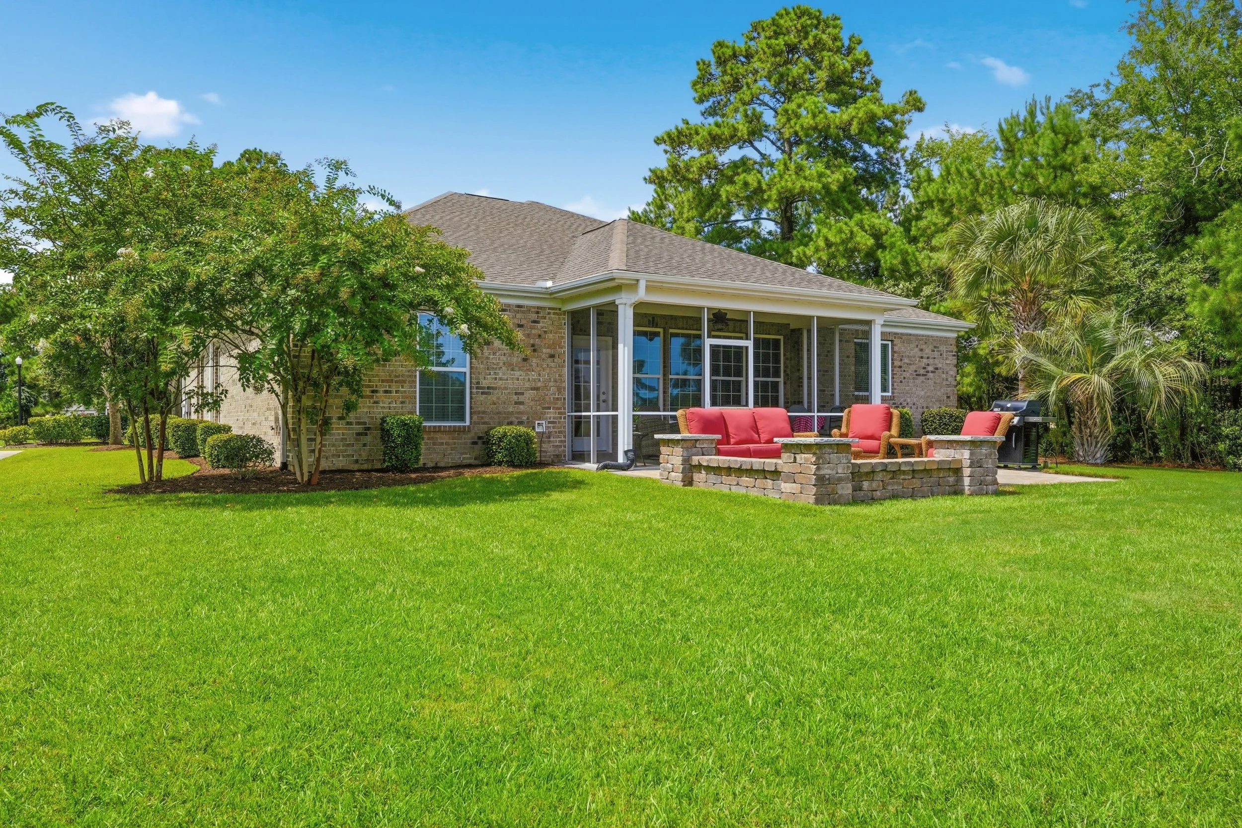 A brick house with a screened porch, surrounded by green grass, golf course, and various trees, with outdoor furniture including a red-cushioned sofa and chairs, and a barbecue grill on a sunny day.
